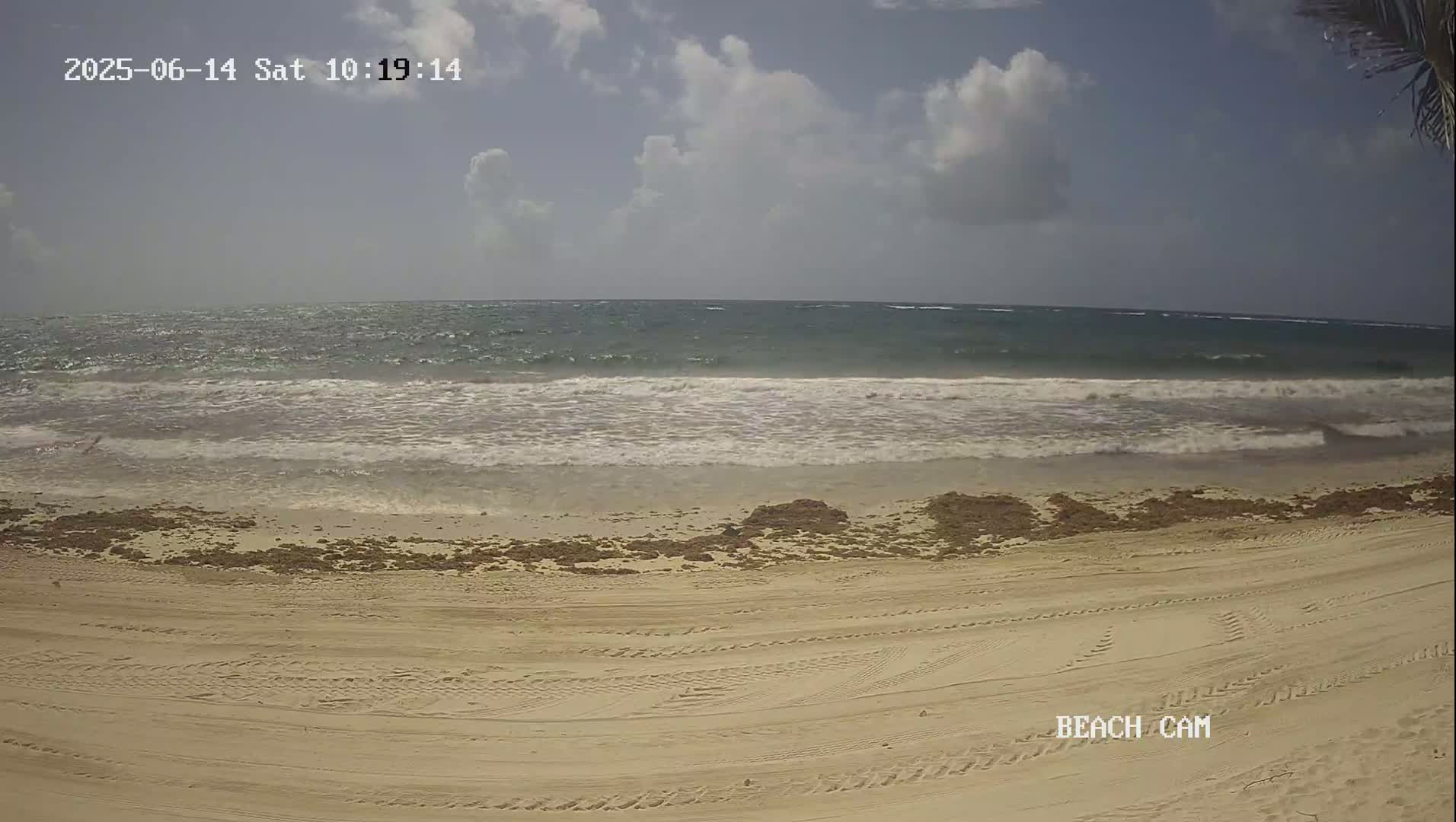 A sandy beach with tire tracks stretches towards a wavy ocean under a partly cloudy sky.