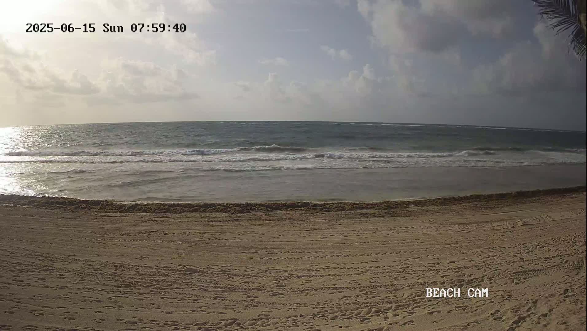A sandy beach with tire tracks and footprints stretches to a calm ocean with small waves under a partly cloudy sky.