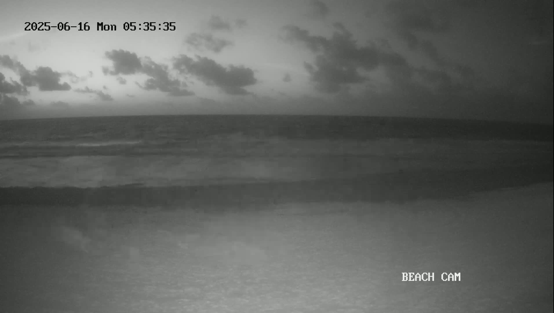 A dark, cloudy pre-dawn sky hangs over a mostly calm ocean meeting a sandy beach.