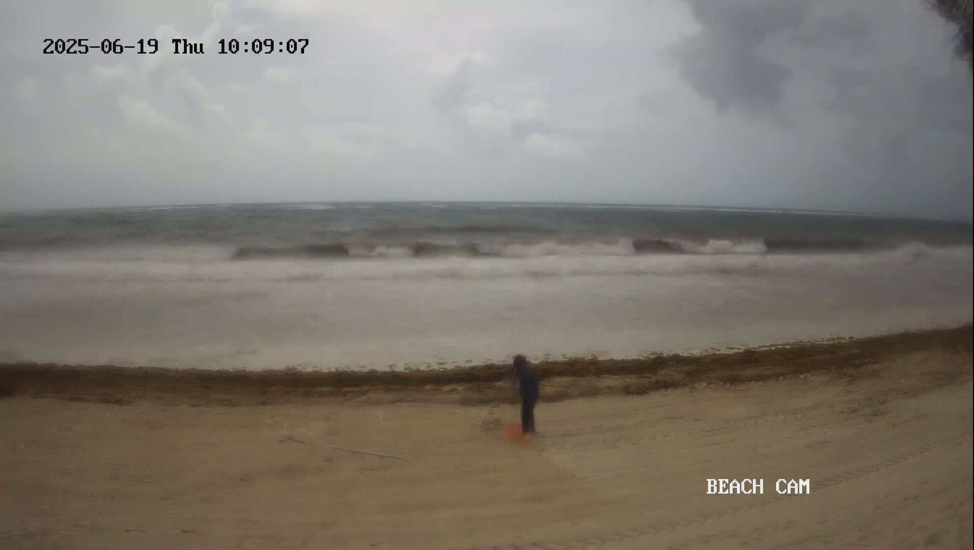 A person stands on a sandy beach with moderate waves crashing on the shore under an overcast sky.