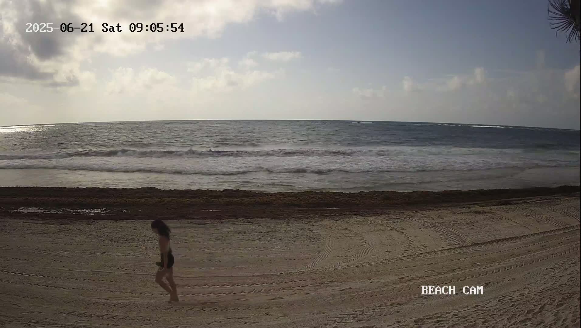 A woman walks along a sandy beach under a partly cloudy sky, toward a somewhat choppy ocean.
