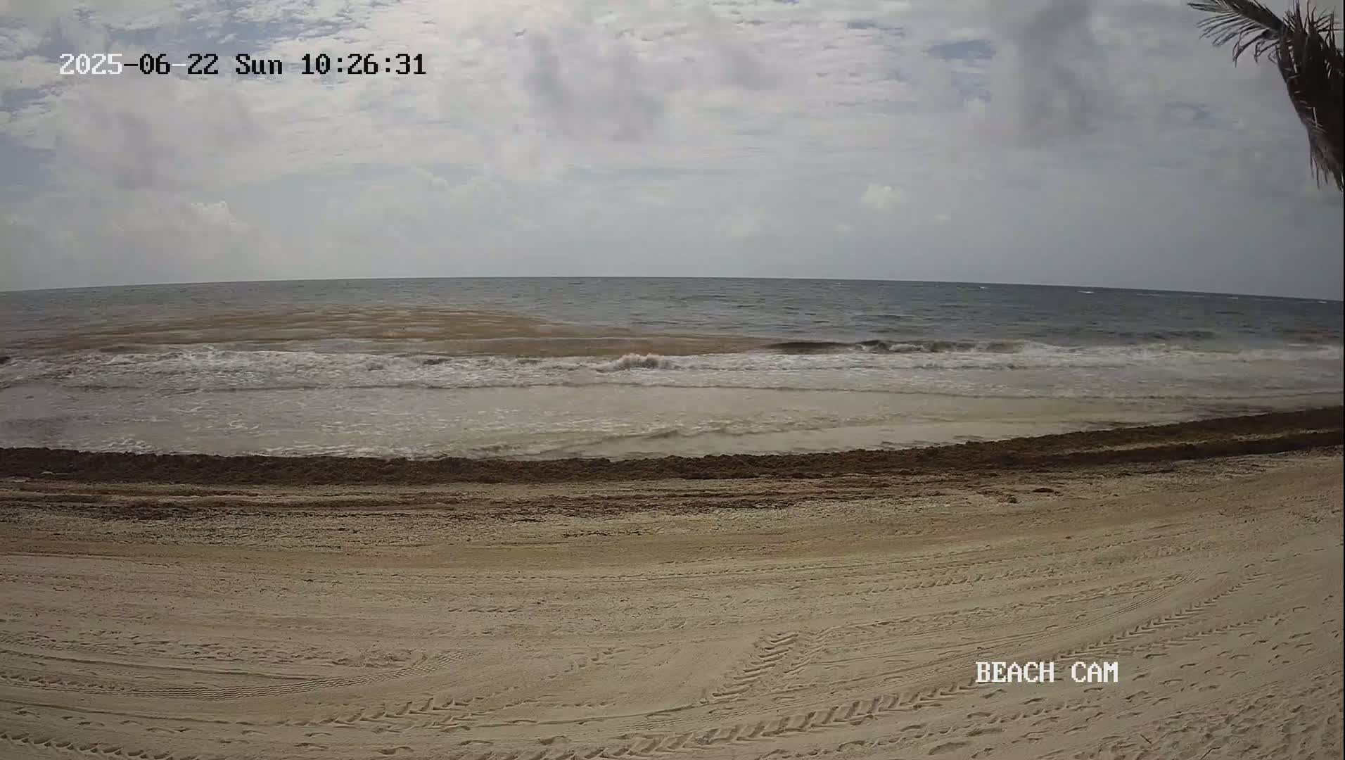 A sandy beach with tire tracks shows gentle waves rolling onto the shore under a partly cloudy sky.