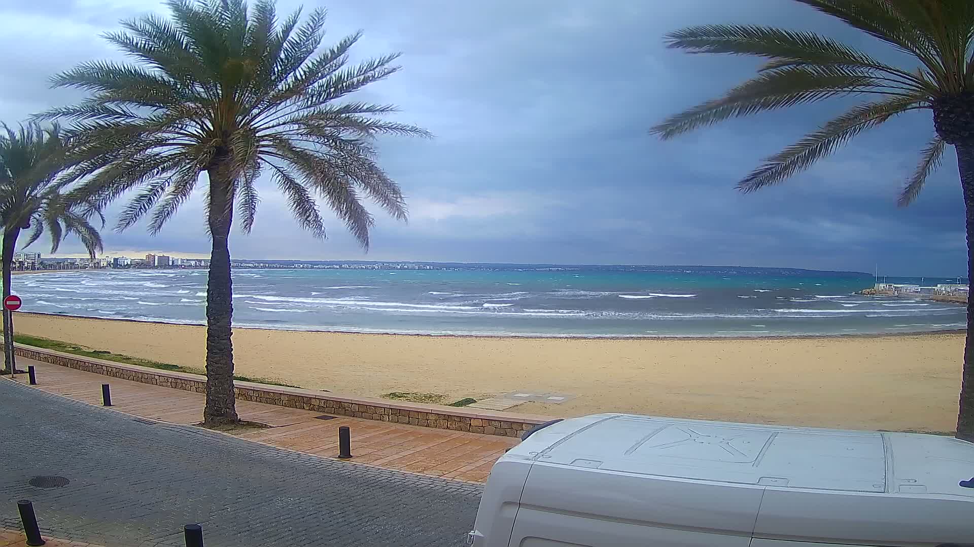 A sandy beach with choppy waves under a cloudy sky is viewed from behind a white van, framed by palm trees and a paved walkway.