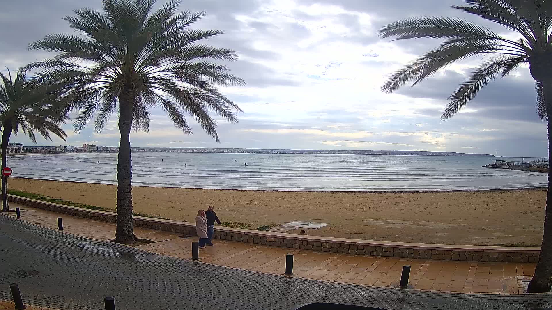 A couple walks along a paved beachfront path beside a sandy beach with calm waves under a cloudy sky.