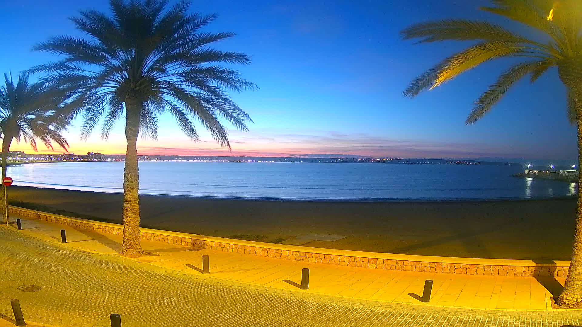 A calm ocean beach at twilight, under a clear blue sky transitioning to pink and orange near the horizon, is bordered by a paved walkway and palm trees.