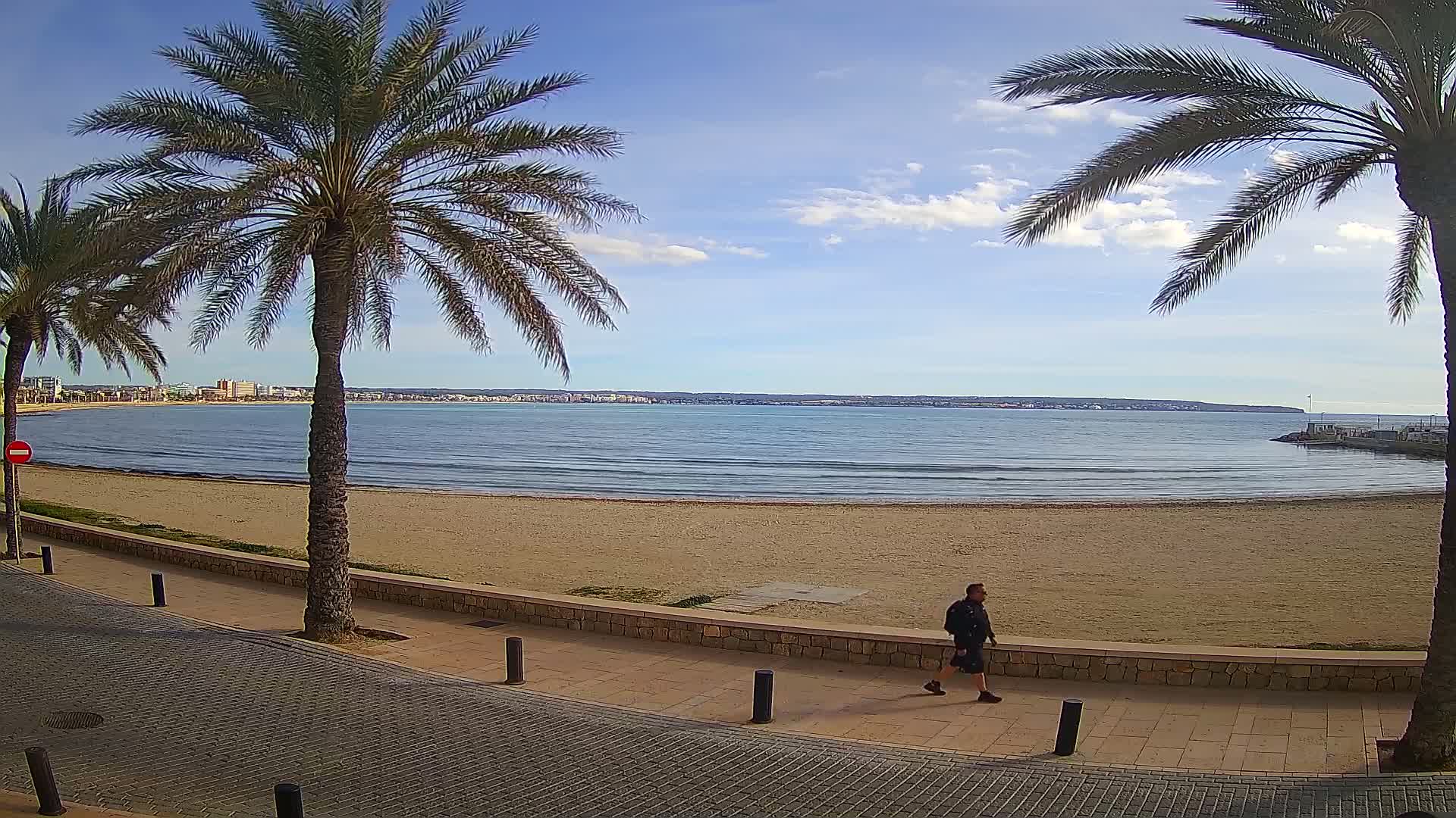 A man walks along a paved beachfront path next to a sandy beach and calm ocean under a mostly sunny sky.