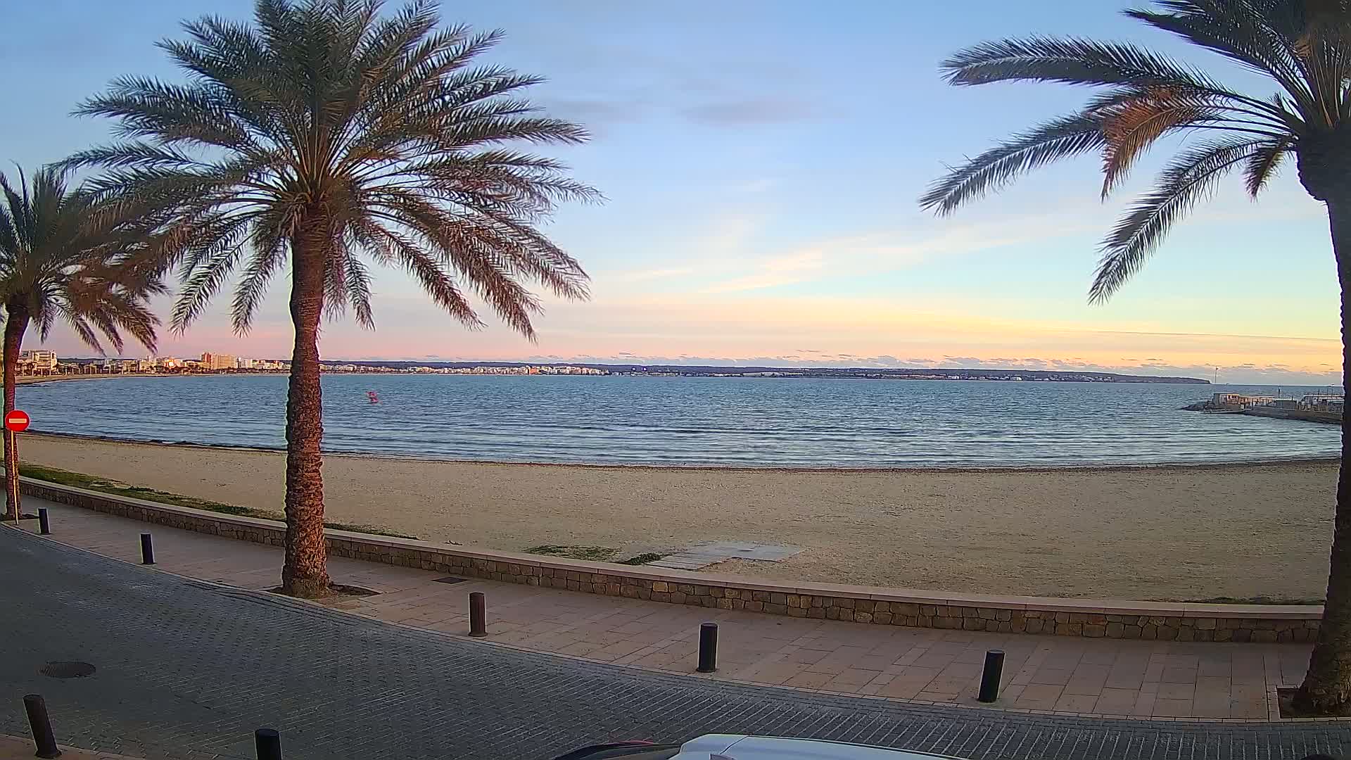A sandy beach and calm sea are seen at sunset, framed by two palm trees and a paved walkway.