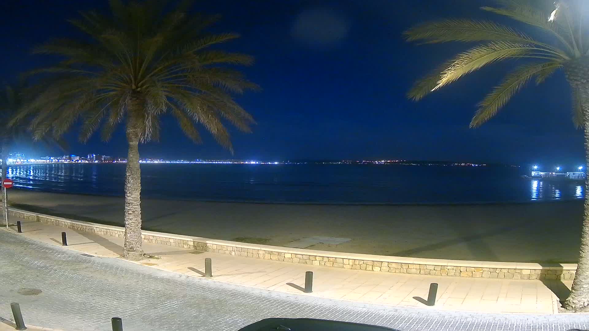 A paved walkway borders a sandy beach at night, overlooking a calm sea with a city skyline faintly illuminated in the distance under a dark blue sky.