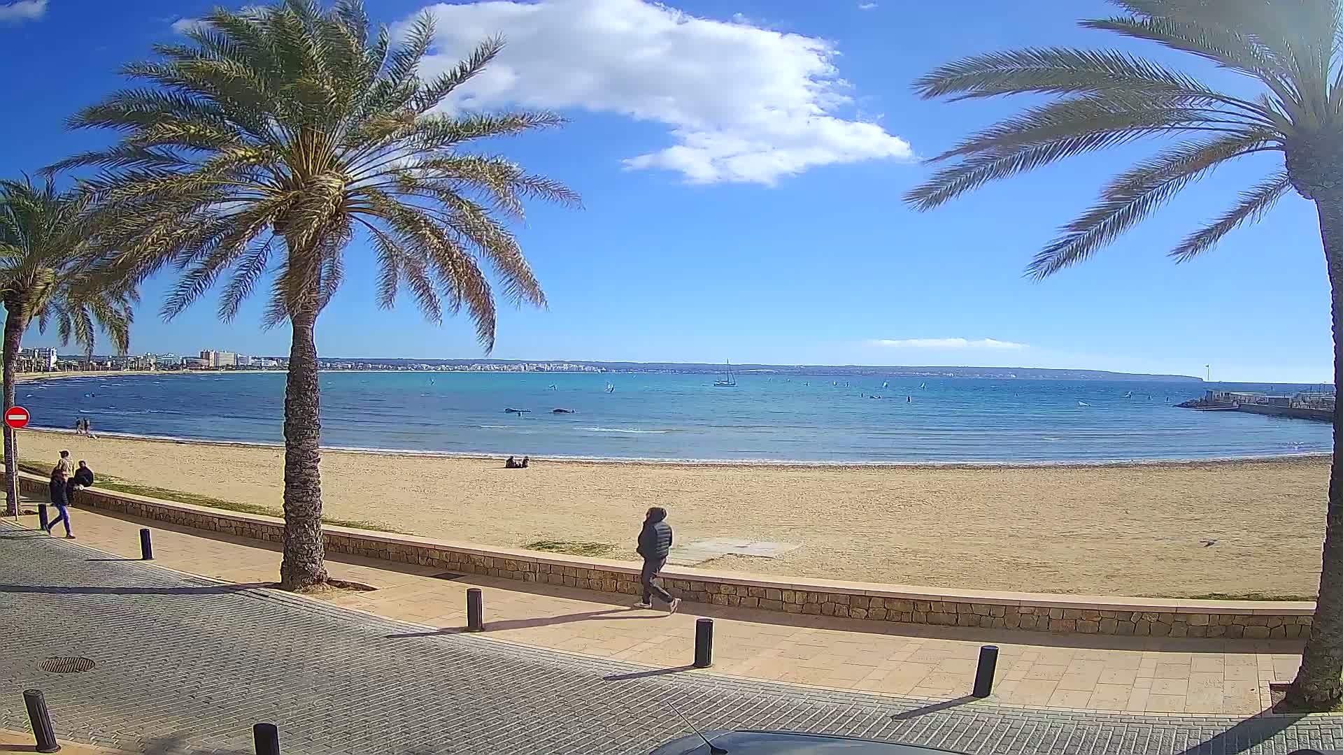 A sandy beach with calm, blue water, palm trees lining a paved walkway, and several people walking on the beach and boardwalk under a mostly sunny sky with a few clouds.