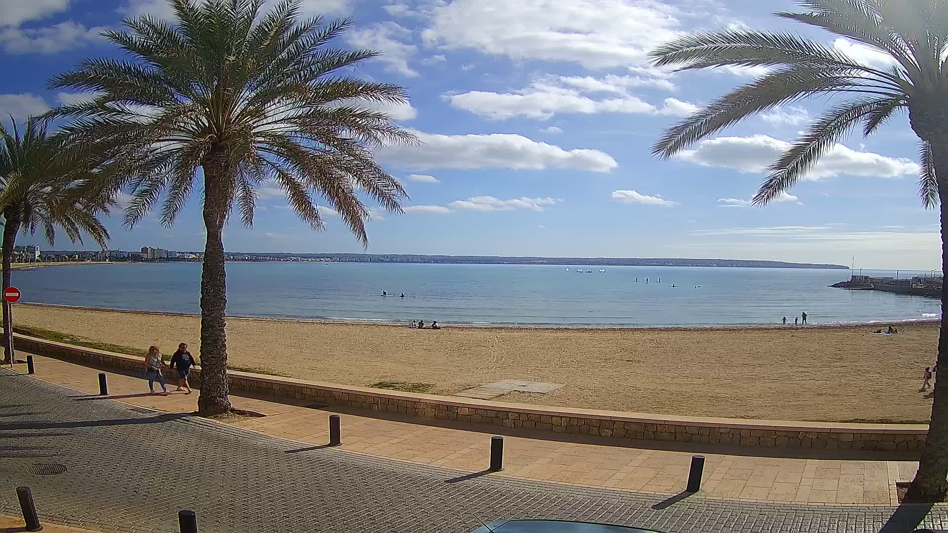 A sandy beach with calm blue water, a few people, and palm trees lining a paved walkway under a partly sunny sky.