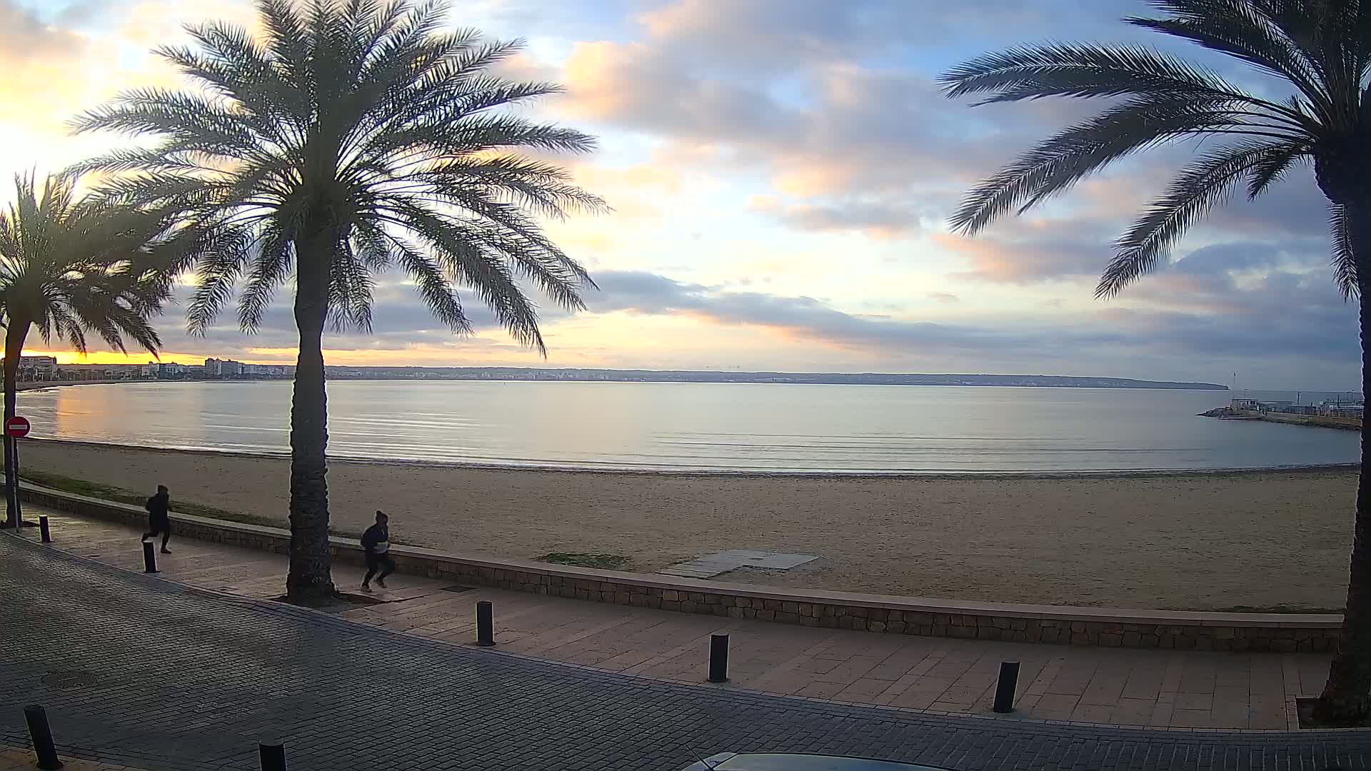 Two people jog along a paved walkway next to a sandy beach at sunrise, with calm water and palm trees in the foreground.