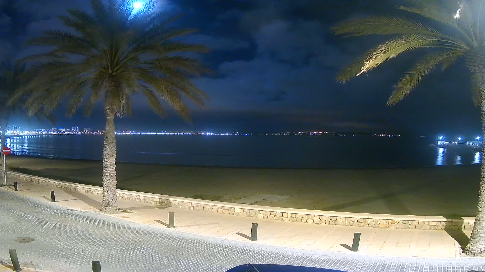 A night scene shows a mostly empty beach with palm trees lining a paved walkway, overlooking a calm, dark-blue sea and a distant, illuminated city skyline under a partly cloudy sky.