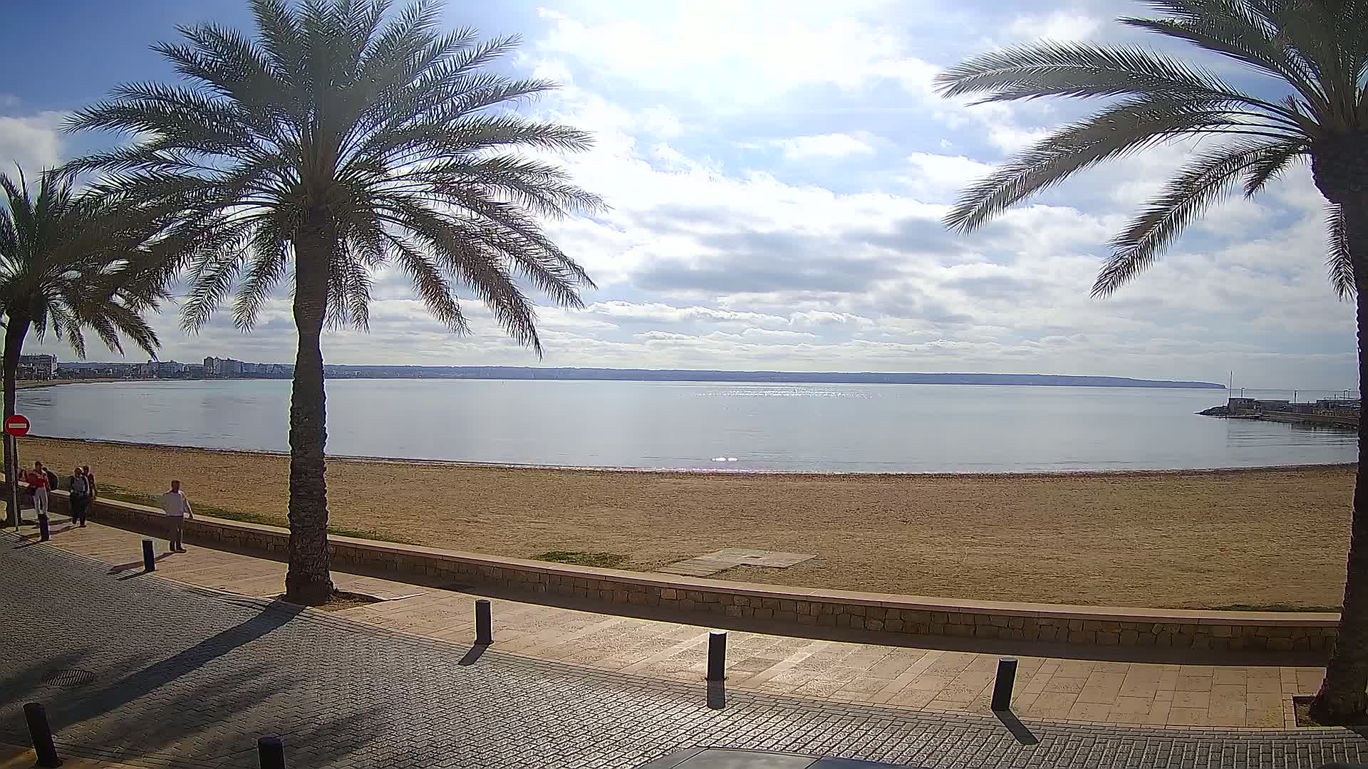 A partly cloudy day shows a sandy beach with calm water, palm trees, and a paved walkway with several people walking.