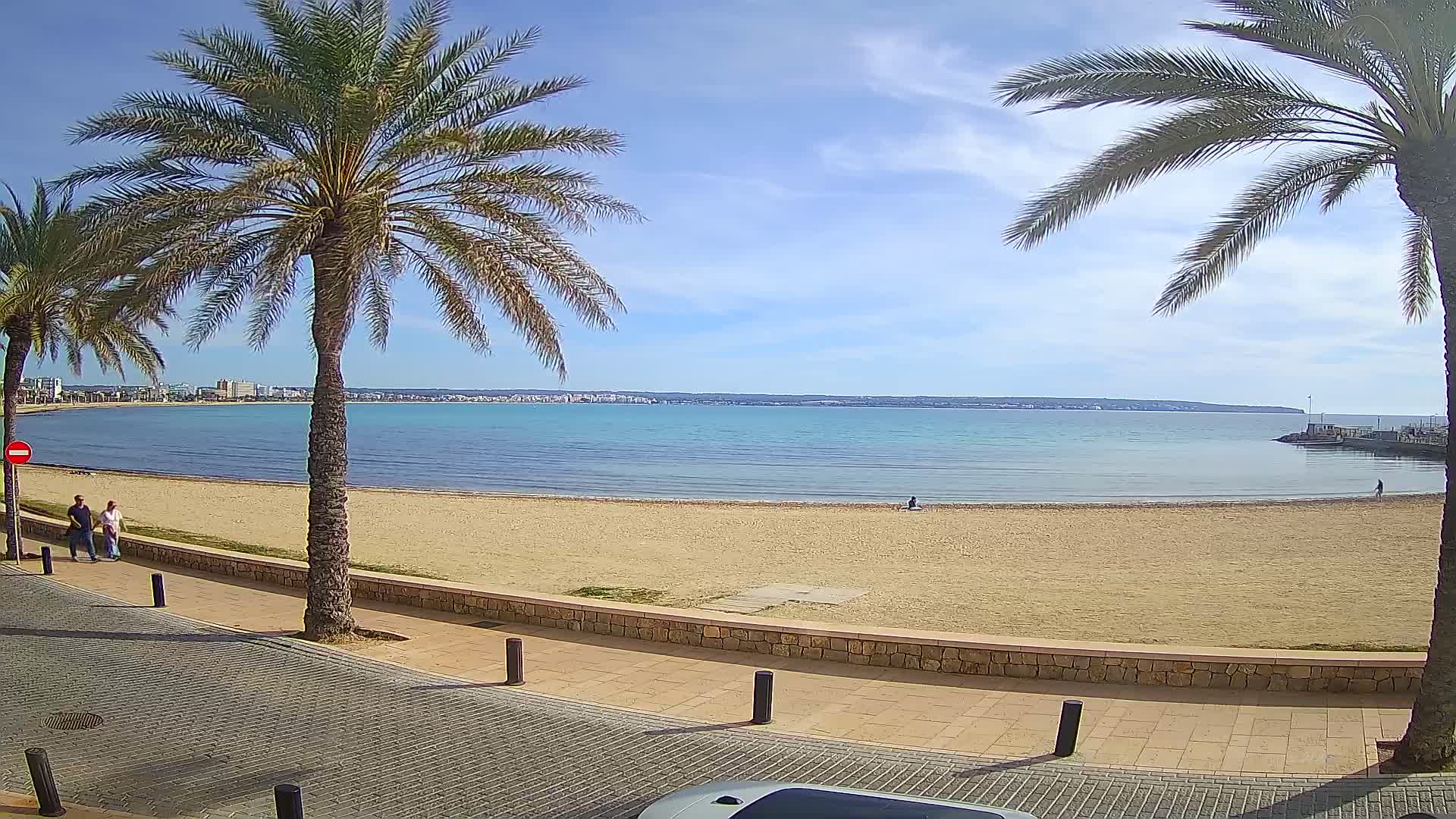 A sandy beach with calm, light-blue water under a mostly sunny sky, flanked by palm trees and a paved walkway with a few people walking.