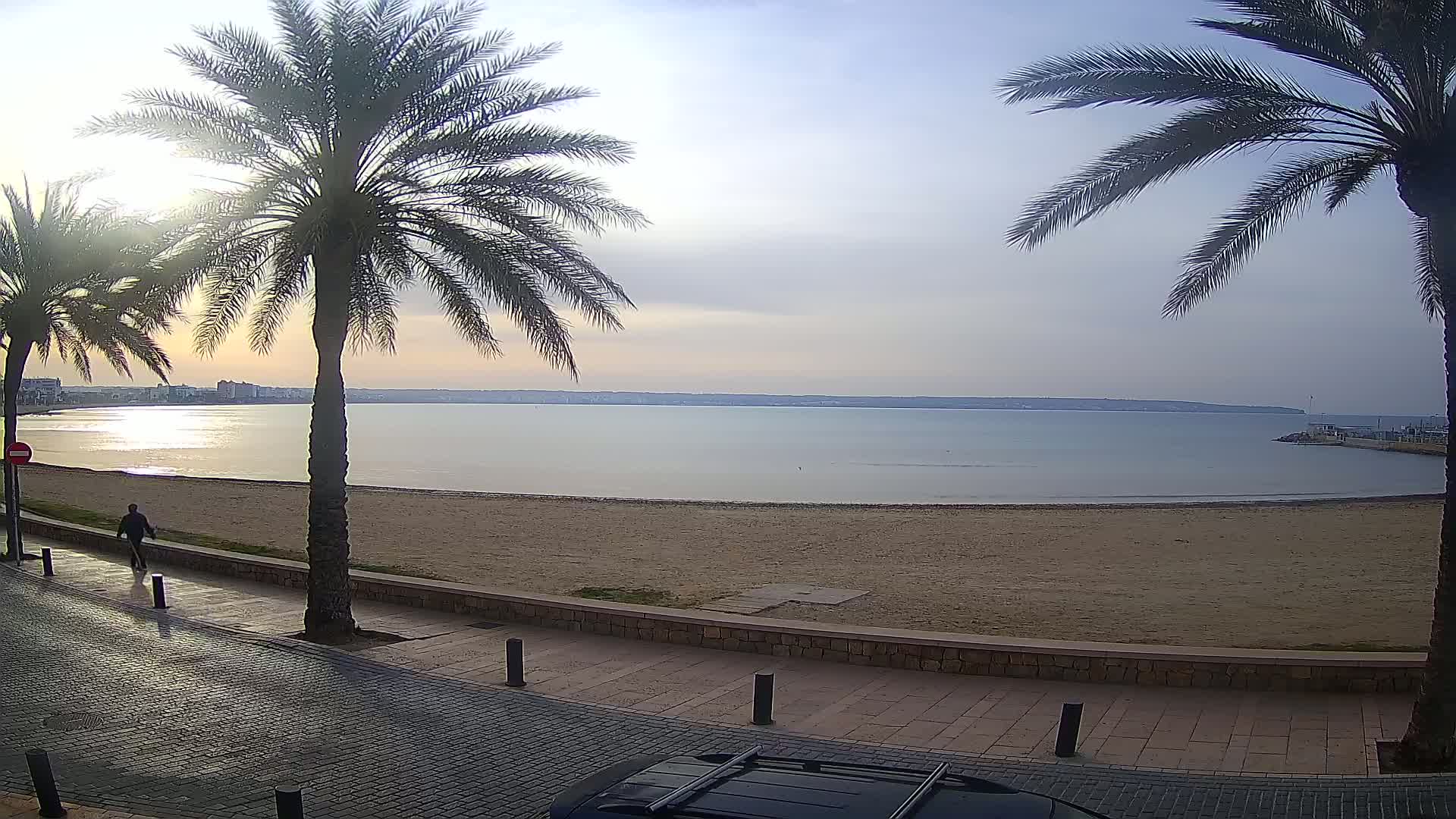 A person walks along a paved path next to a sandy beach and calm sea, under a mostly clear, bright sky.