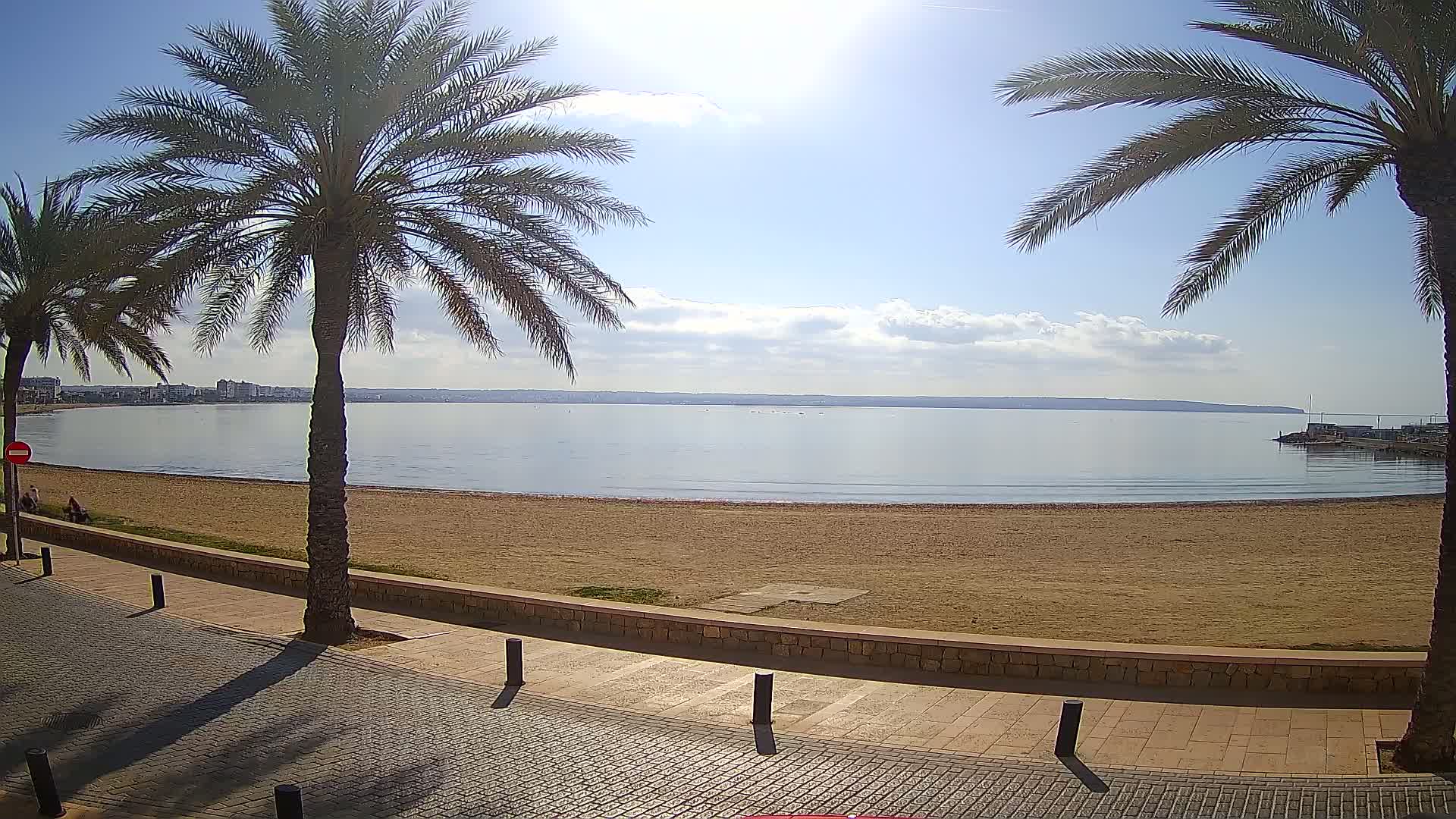 A sandy beach with calm water and a few people, bordered by palm trees and a paved walkway under a mostly sunny sky.