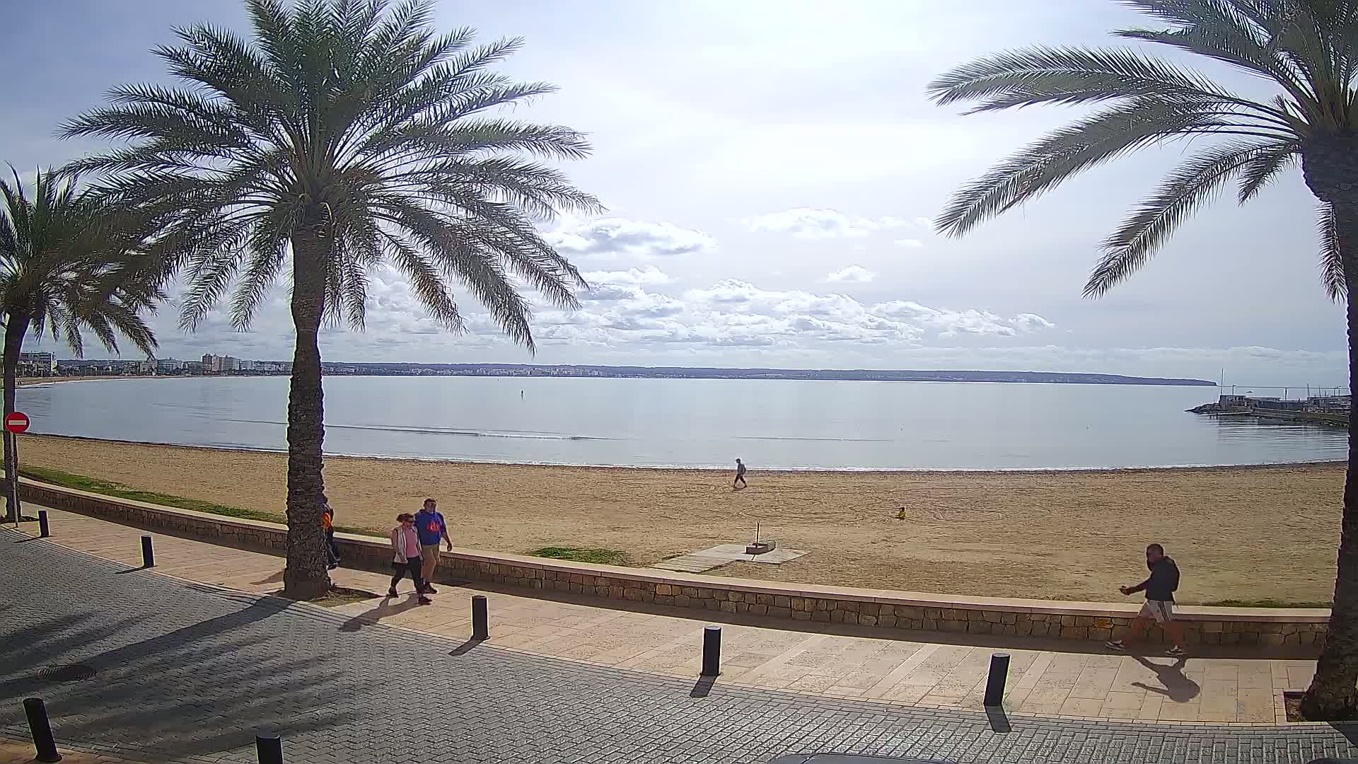 A sandy beach with calm water and a few people walking along a paved walkway, under a partly cloudy sky.
