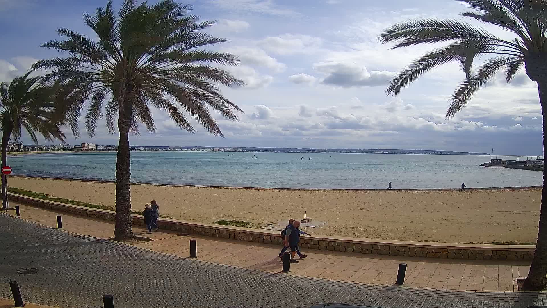 A sandy beach with calm water stretches to a distant shoreline under a partly cloudy sky, framed by palm trees and a paved walkway with a few people strolling.