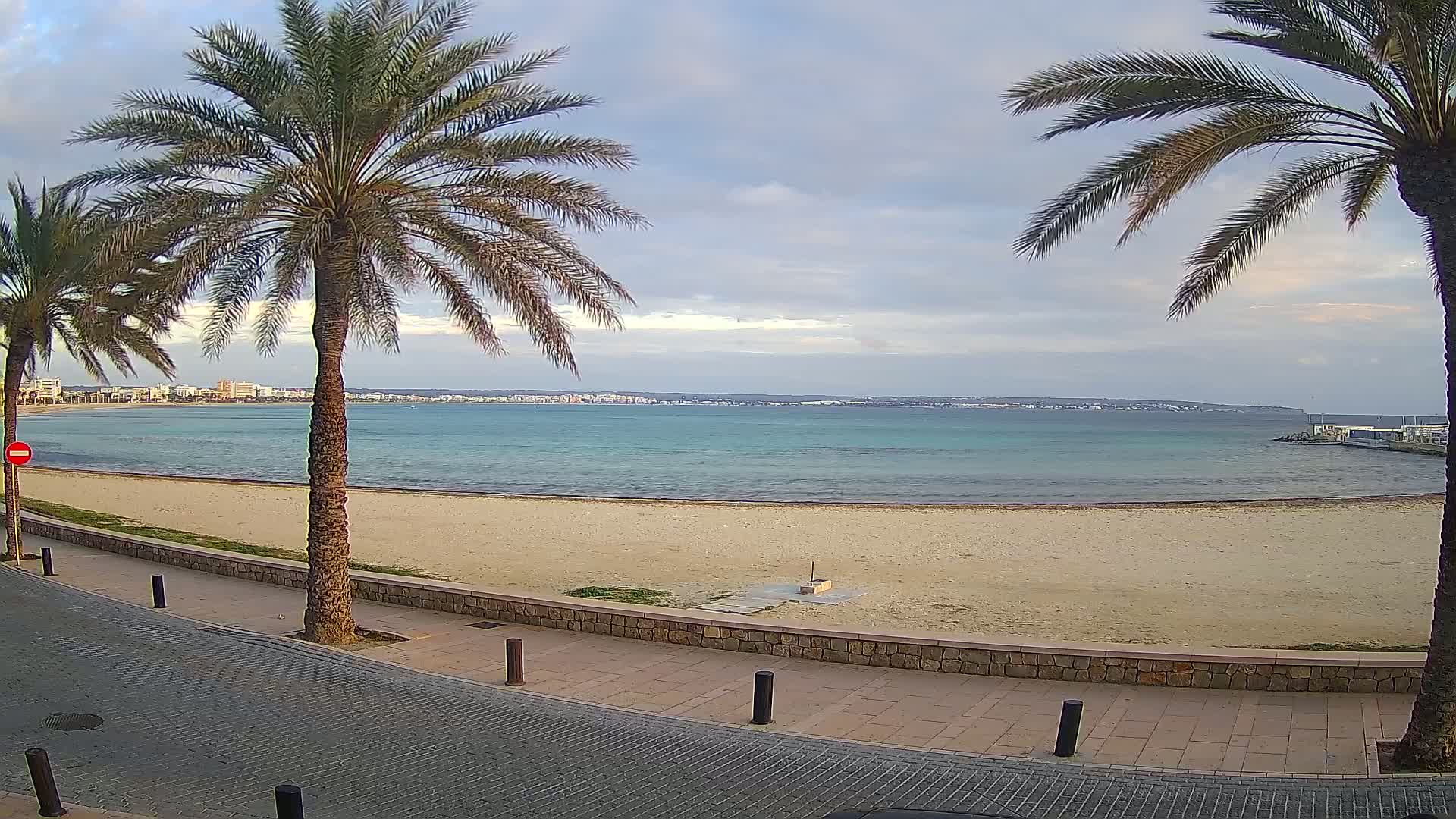 A sandy beach with calm, teal water stretches out under a mostly cloudy sky, flanked by palm trees and a paved walkway.