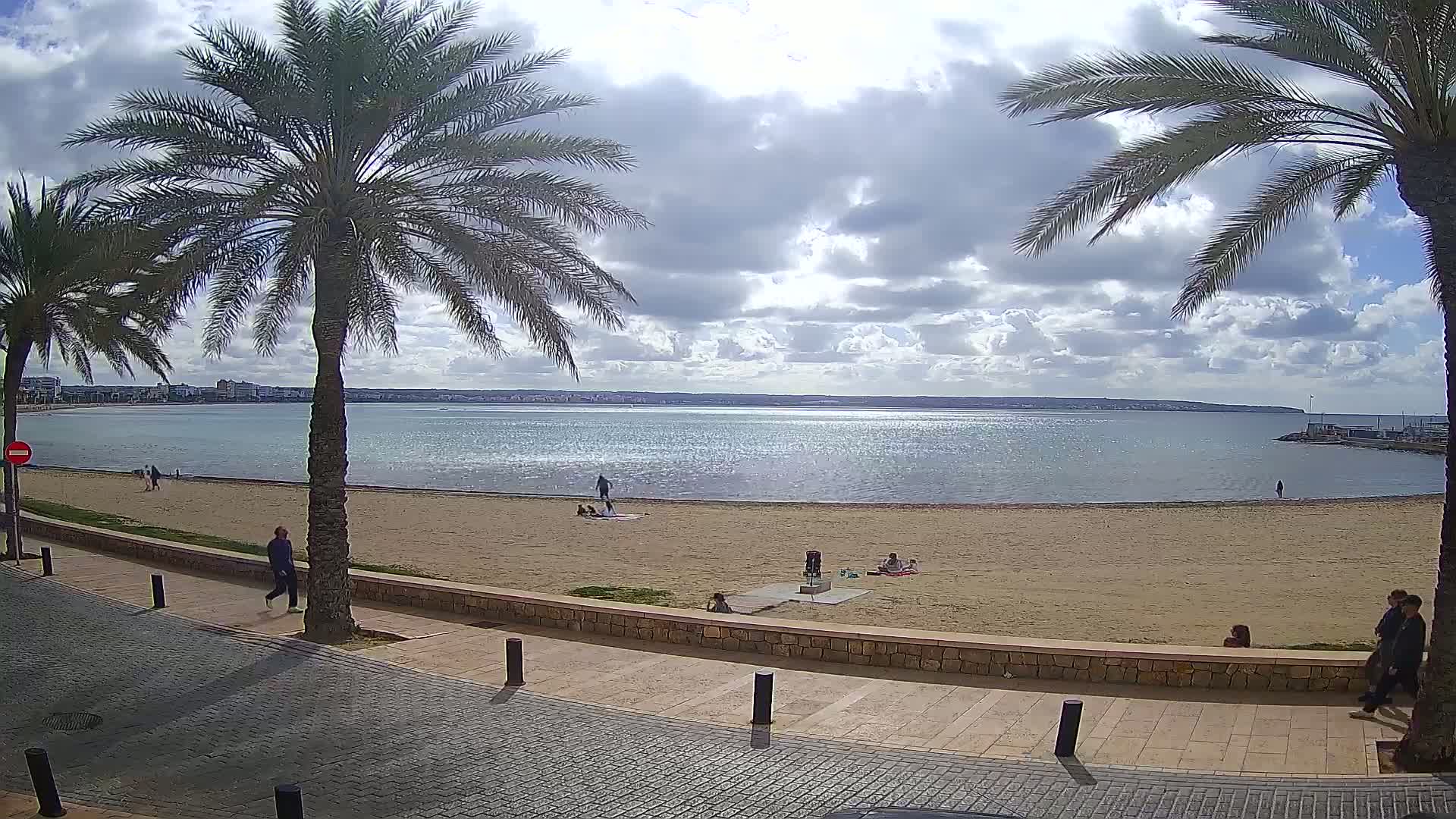 A partly cloudy day at a sandy beach with calm water, palm trees, and a few people walking and sitting.