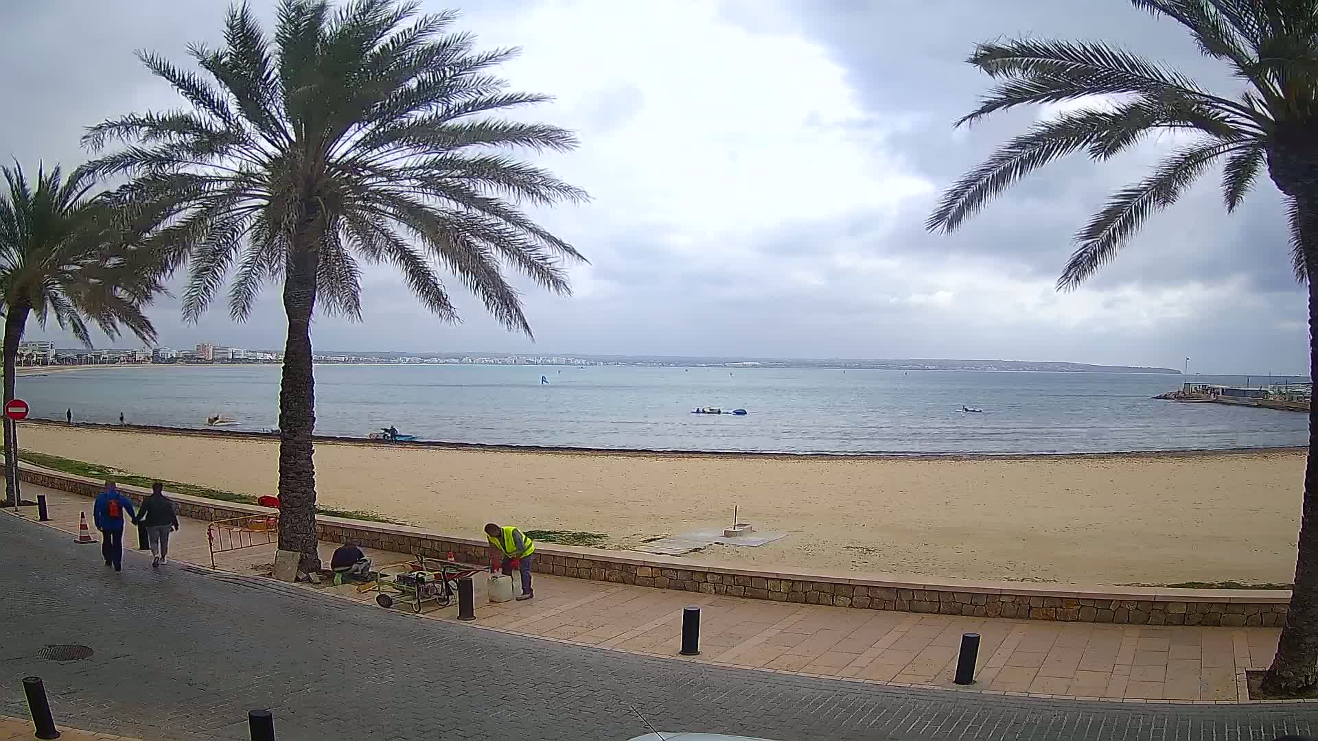 A sandy beach with palm trees lines a paved walkway overlooking calm, gray water under a cloudy sky, with a few people walking on the beach and others working near the walkway.