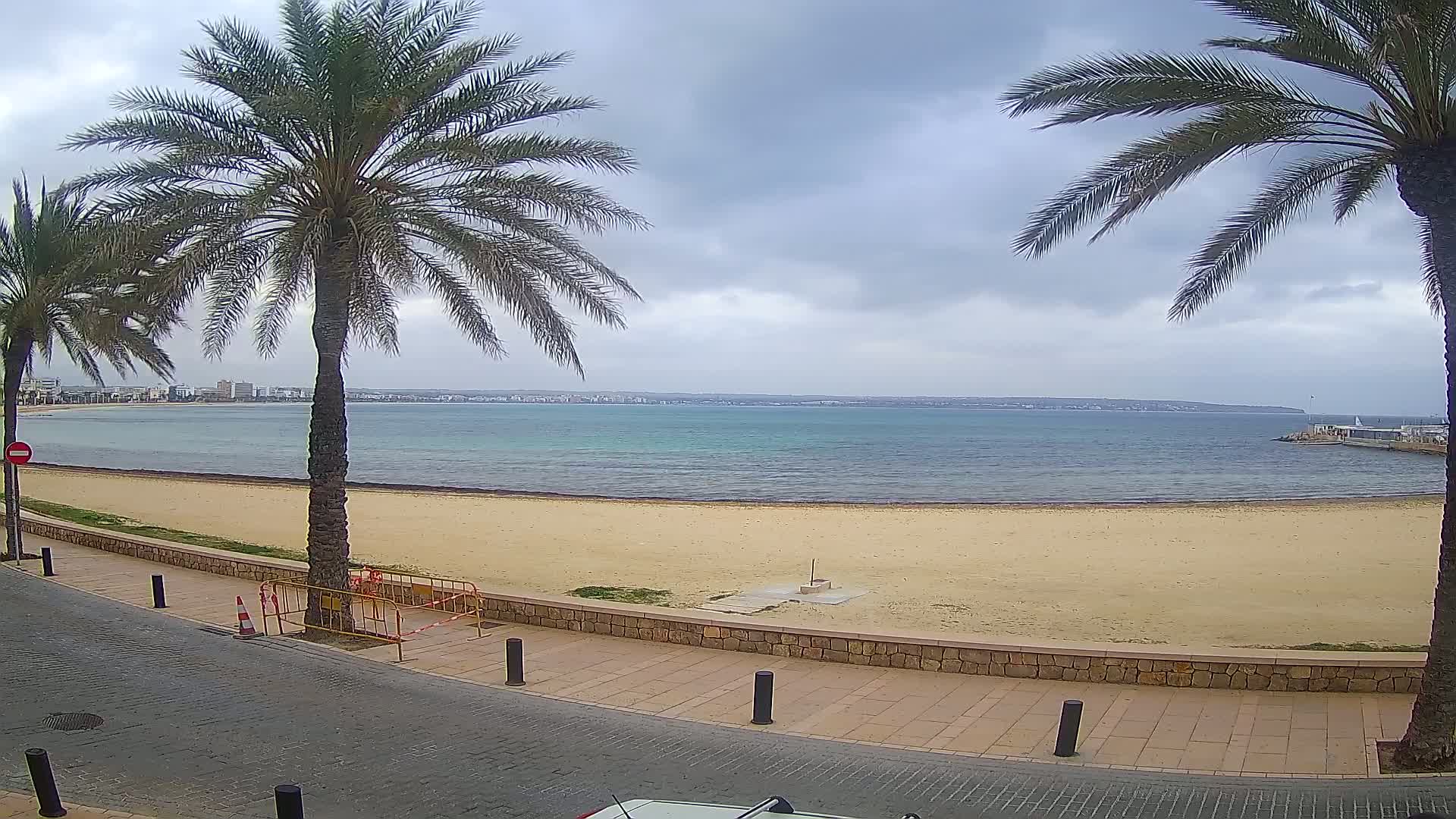 A sandy beach with calm, turquoise water under a cloudy sky is bordered by a paved walkway lined with palm trees.