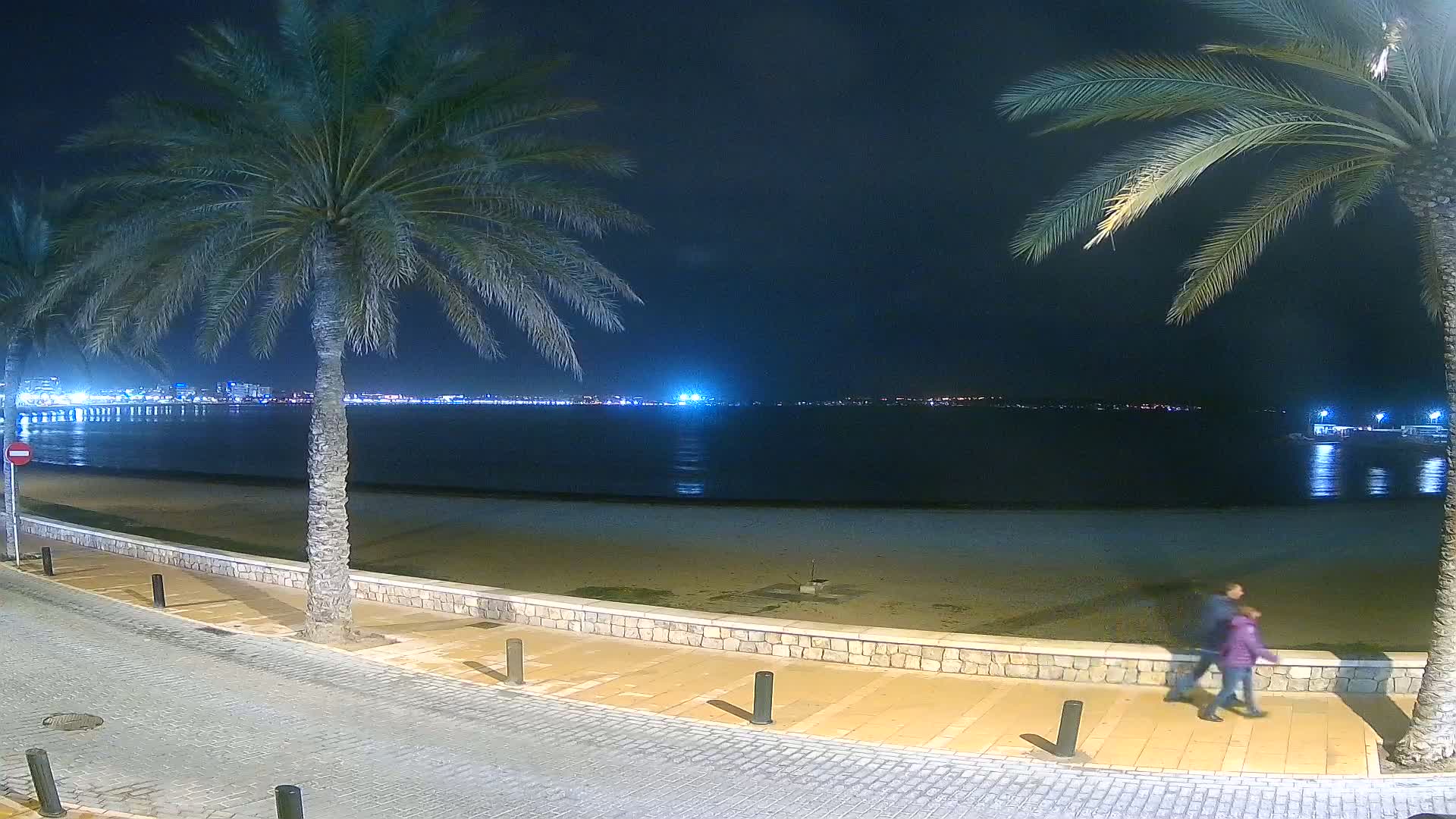 A nighttime beach scene shows two people walking along a paved path next to a sandy beach and calm water, with palm trees lining the path and city lights visible in the distance.