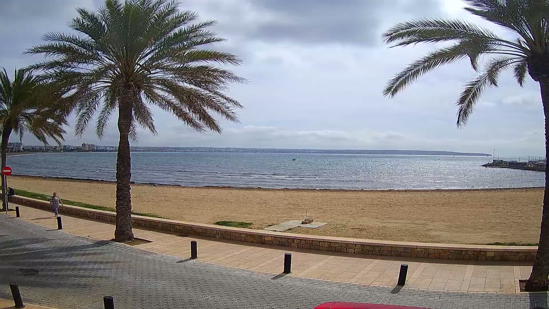 A sandy beach with calm water and palm trees lines a paved walkway under a partly cloudy sky.