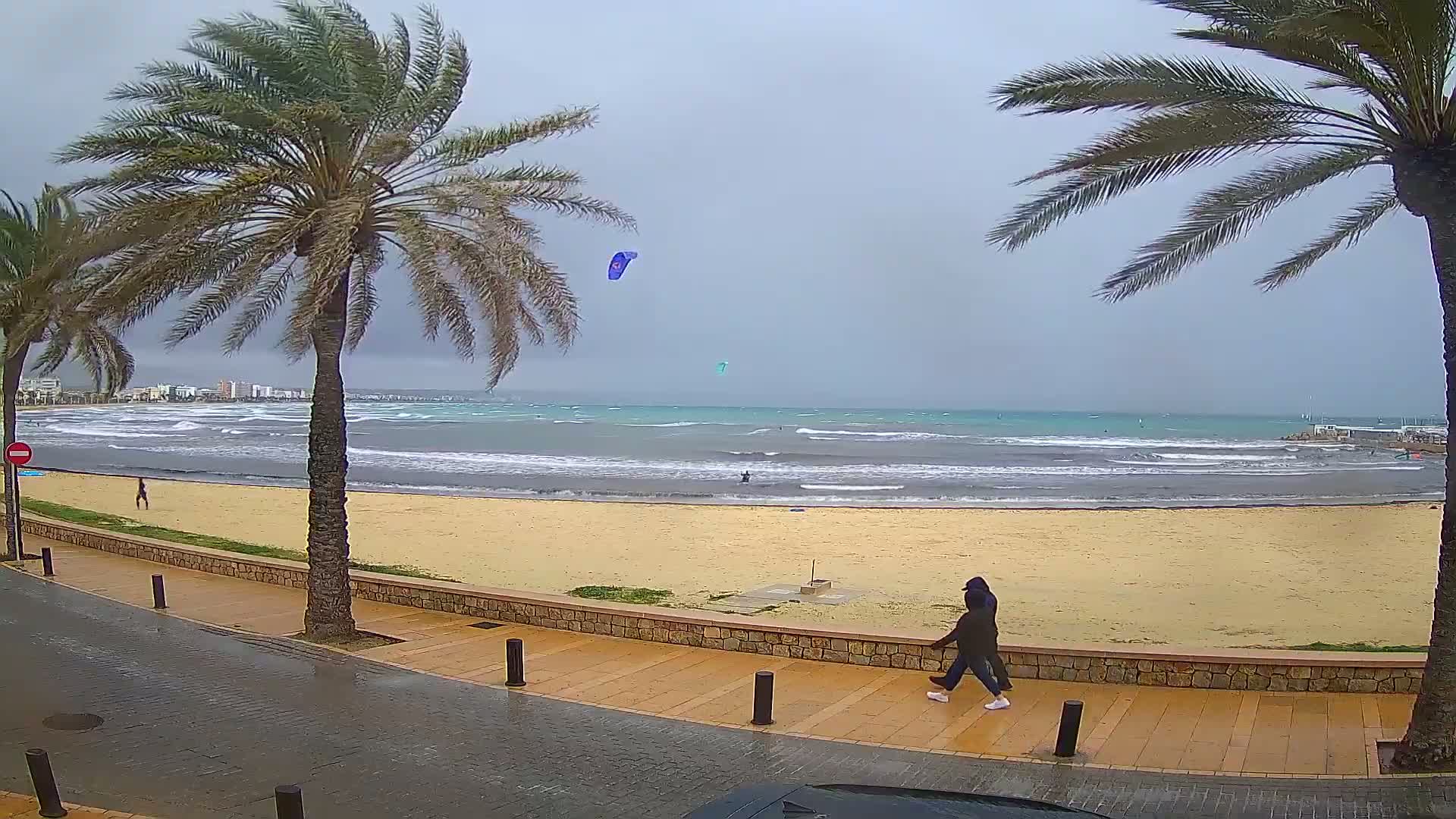A person walks along a paved beachfront path next to a sandy beach with choppy water and a few kite surfers, under an overcast sky.