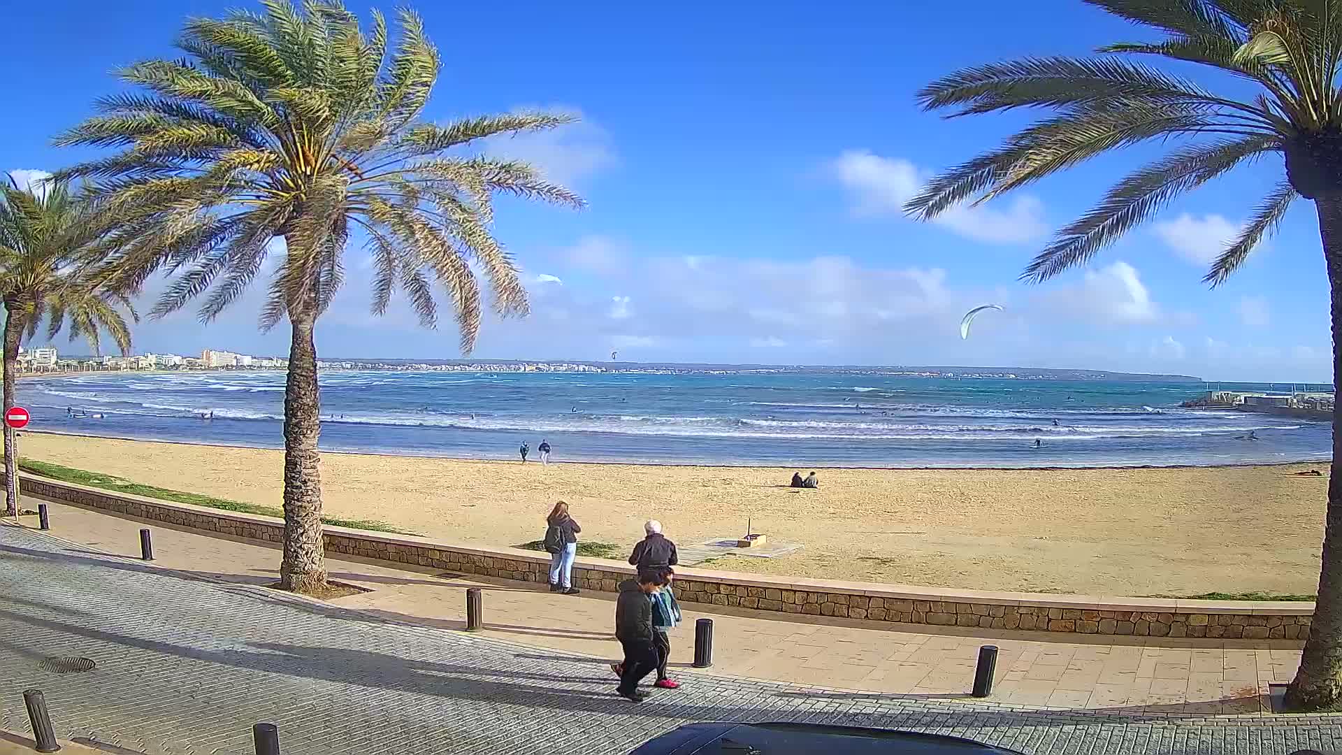 A sandy beach with palm trees and a few people, under a mostly sunny sky with some clouds, shows a calm ocean with some surfers and a kitesurfer in the distance.