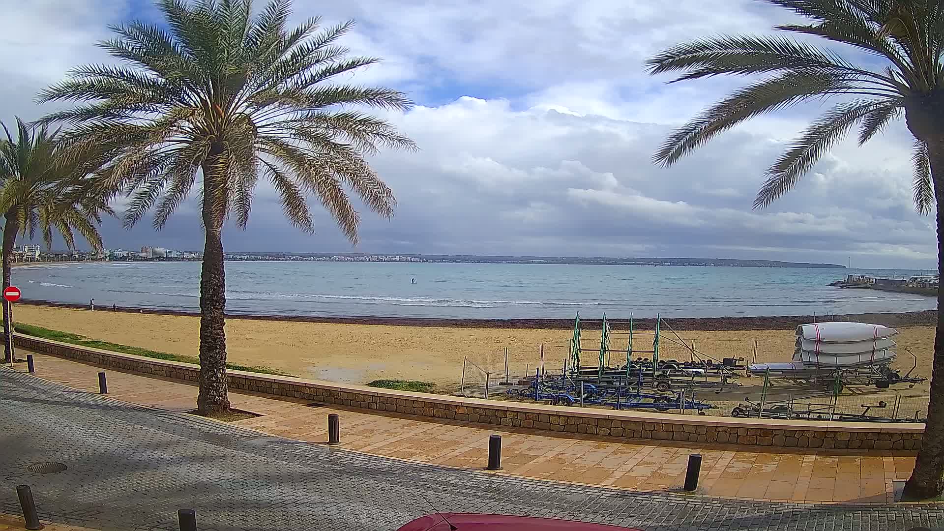 A sandy beach with calm waves and several sailboats on trailers is seen from a paved walkway lined with palm trees under a cloudy sky.