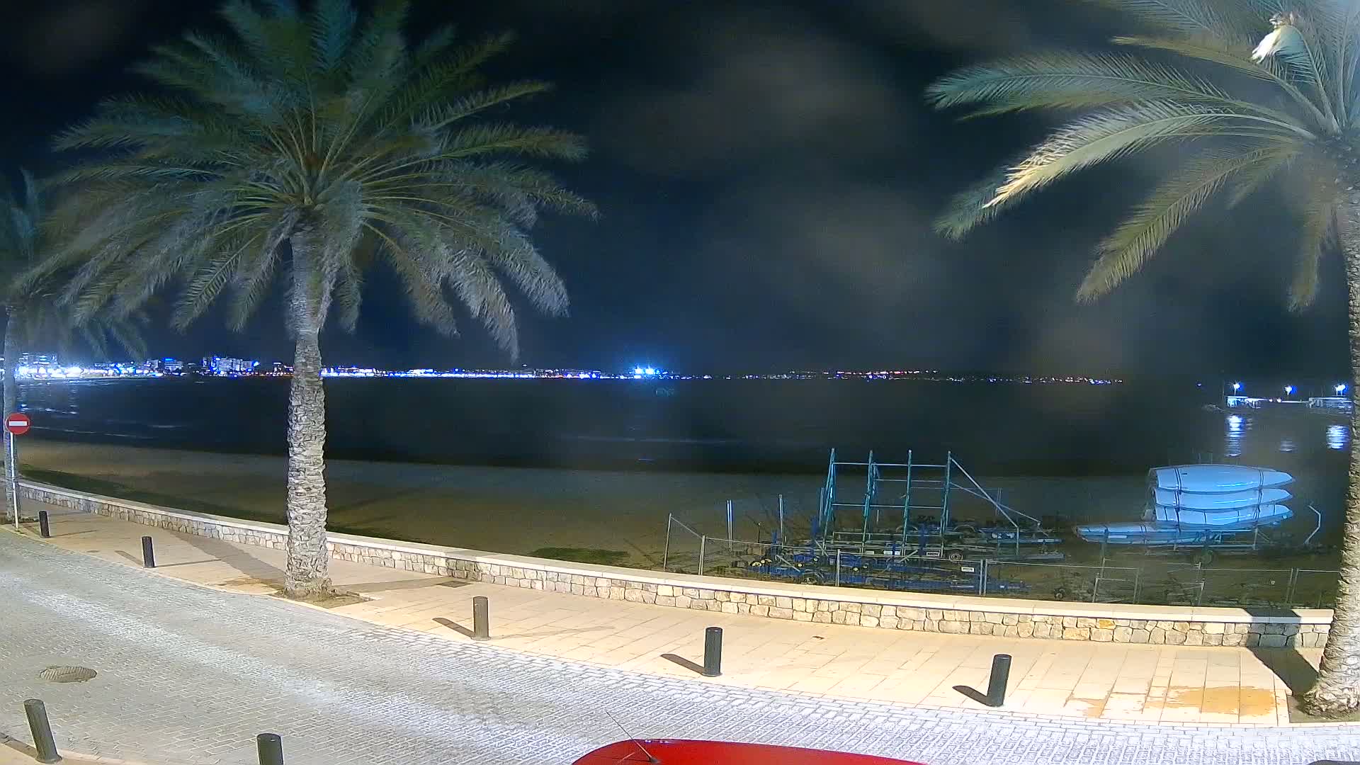 A nighttime view of a beach with palm trees, a calm sea, and several boats stored on the sand near a paved walkway.