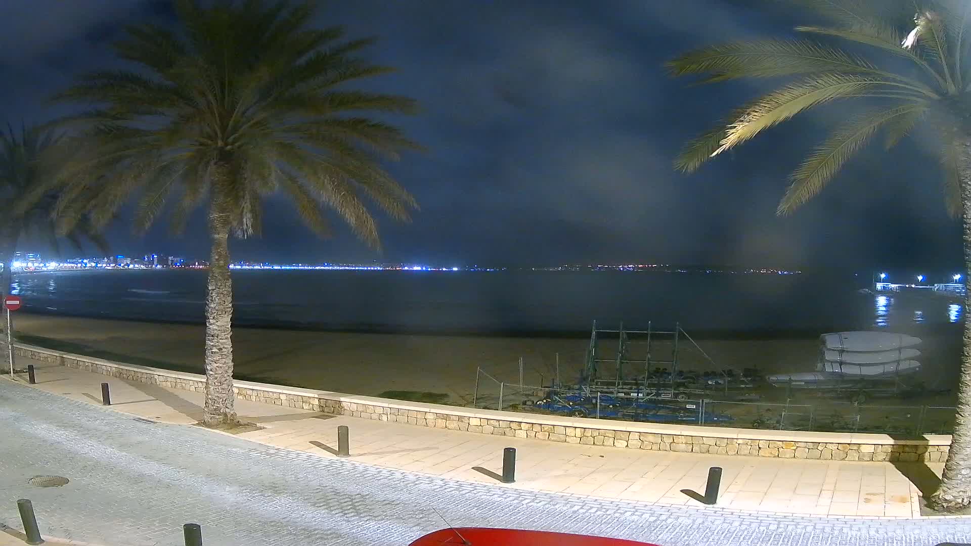 A nighttime view of a calm beach with palm trees, a city skyline in the distance, and several boats resting on the sand.