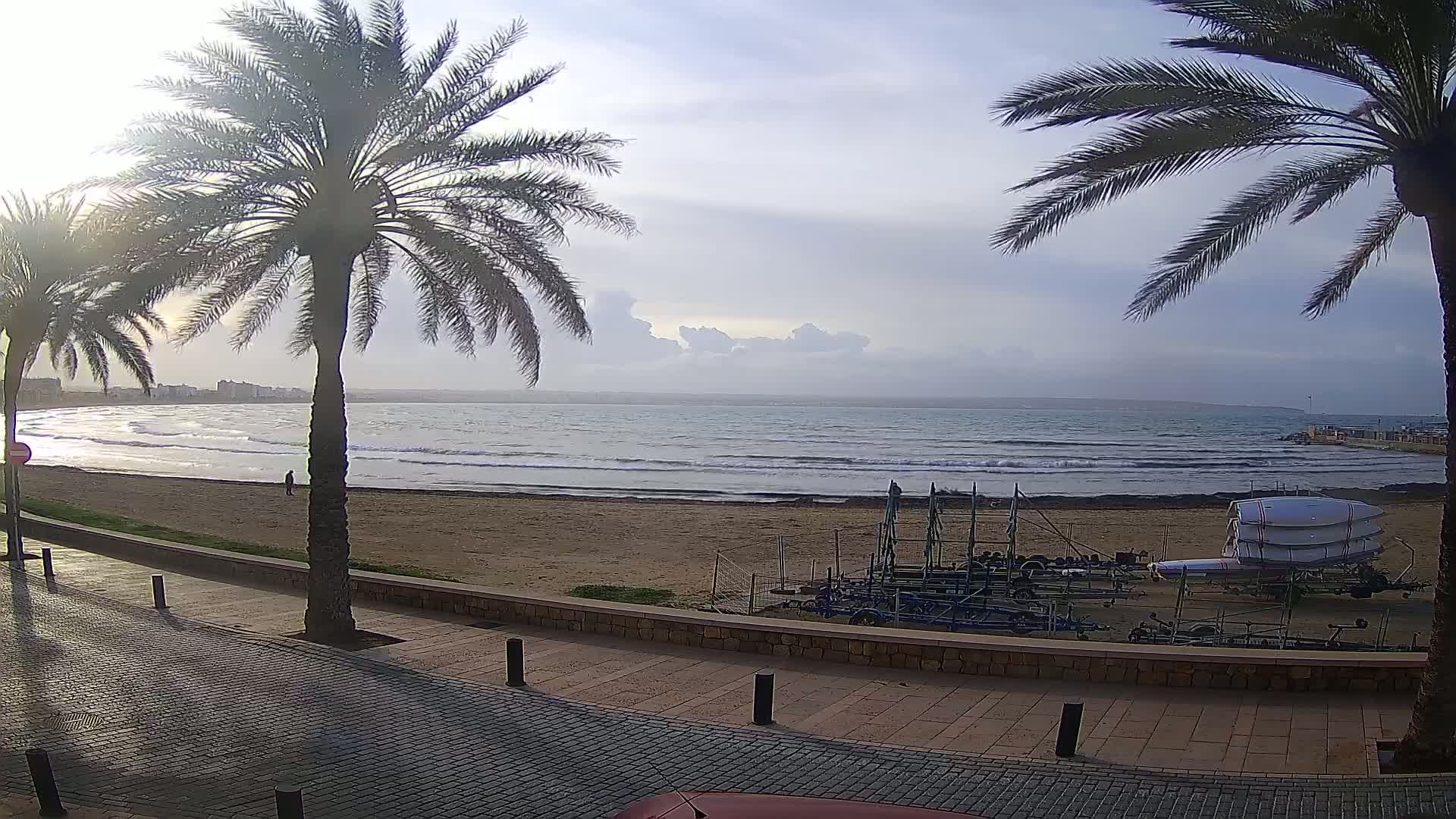 A sandy beach with gentle waves and palm trees lining a paved walkway, under a mostly cloudy sky.