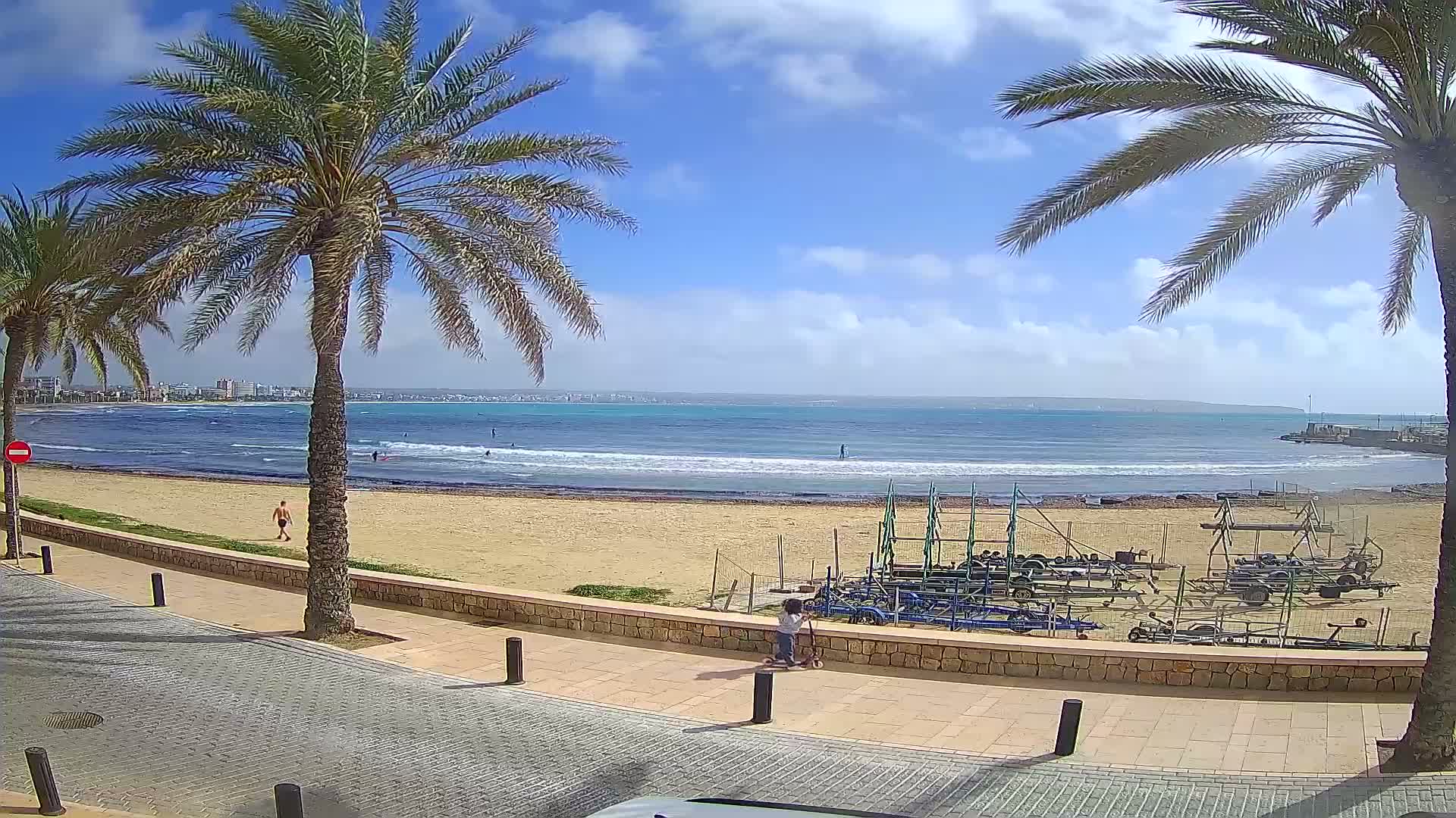 A sandy beach with palm trees lining a paved walkway overlooks a calm ocean with a few people swimming and surfing under a partly cloudy sky.