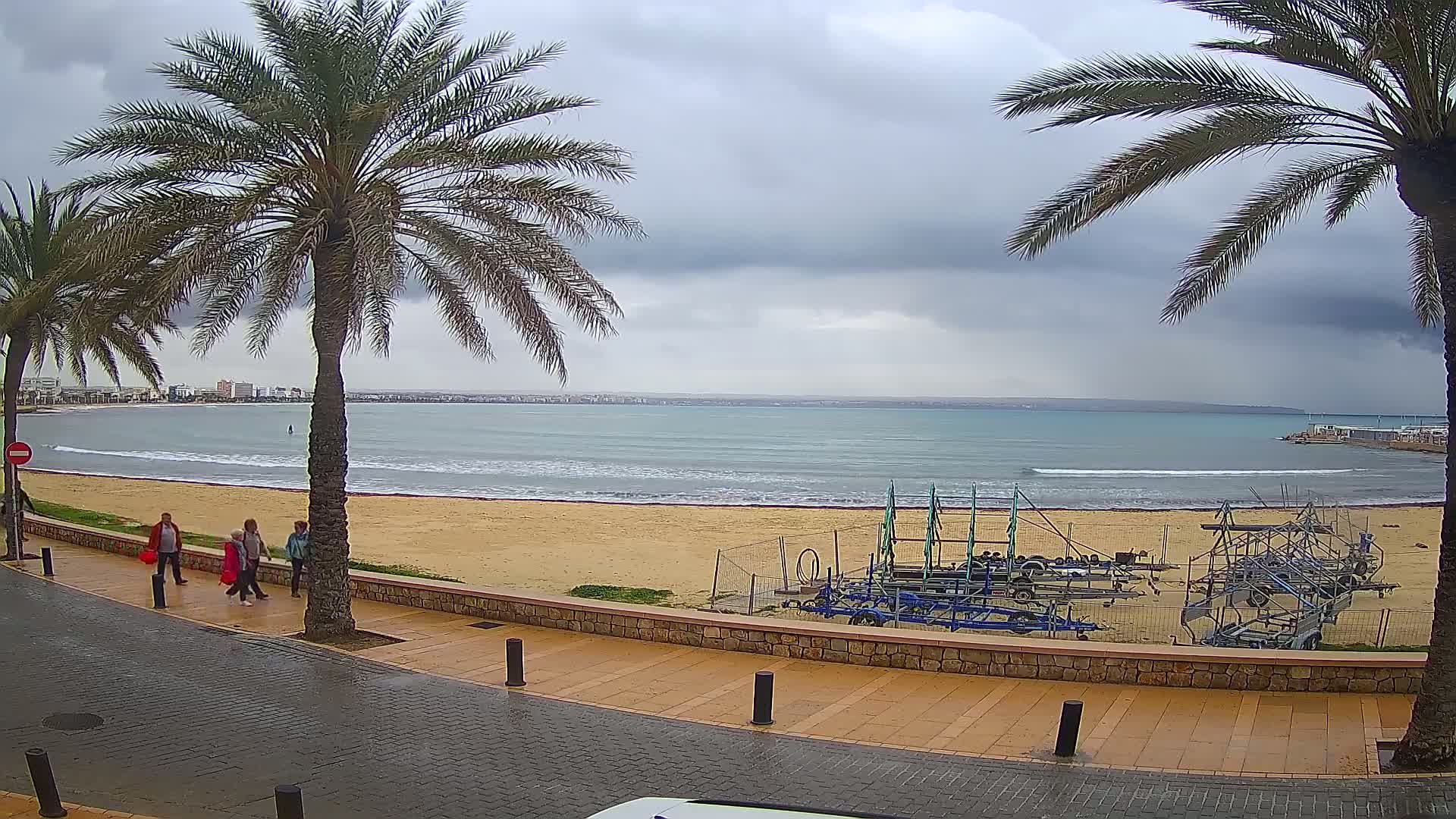 A wet, paved walkway overlooks a sandy beach with calm waves under a cloudy sky, palm trees lining the path, and several people walking along the shore.
