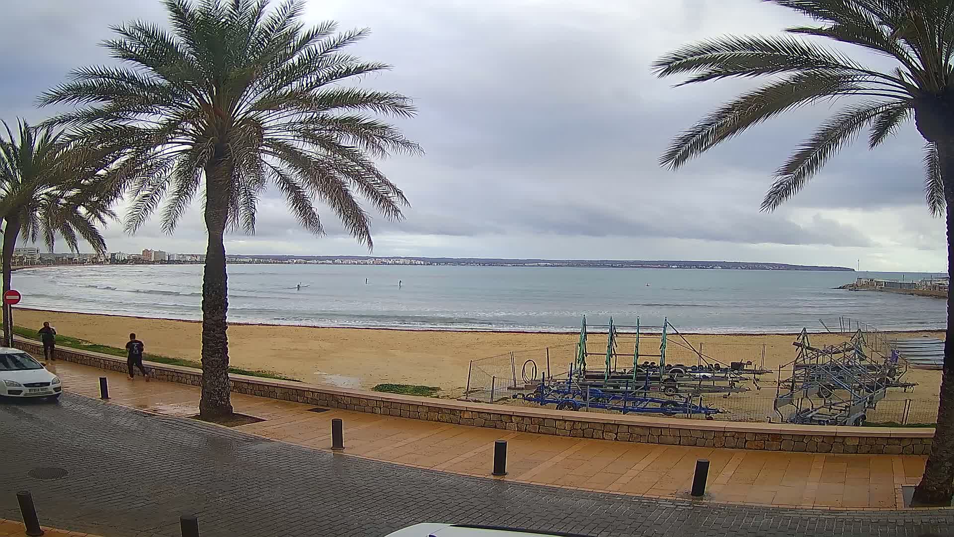 A sandy beach with palm trees lines a paved walkway overlooking a calm sea under a cloudy sky.