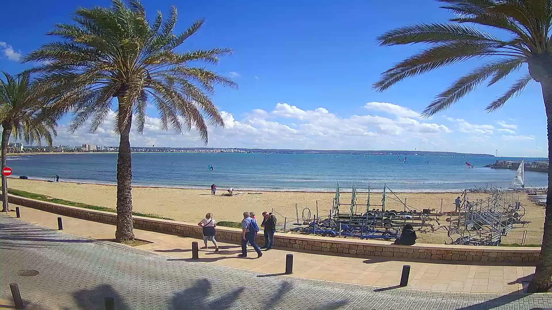 A sandy beach with palm trees lines a paved walkway overlooking a calm, blue ocean with scattered sailboats under a partly cloudy sky.