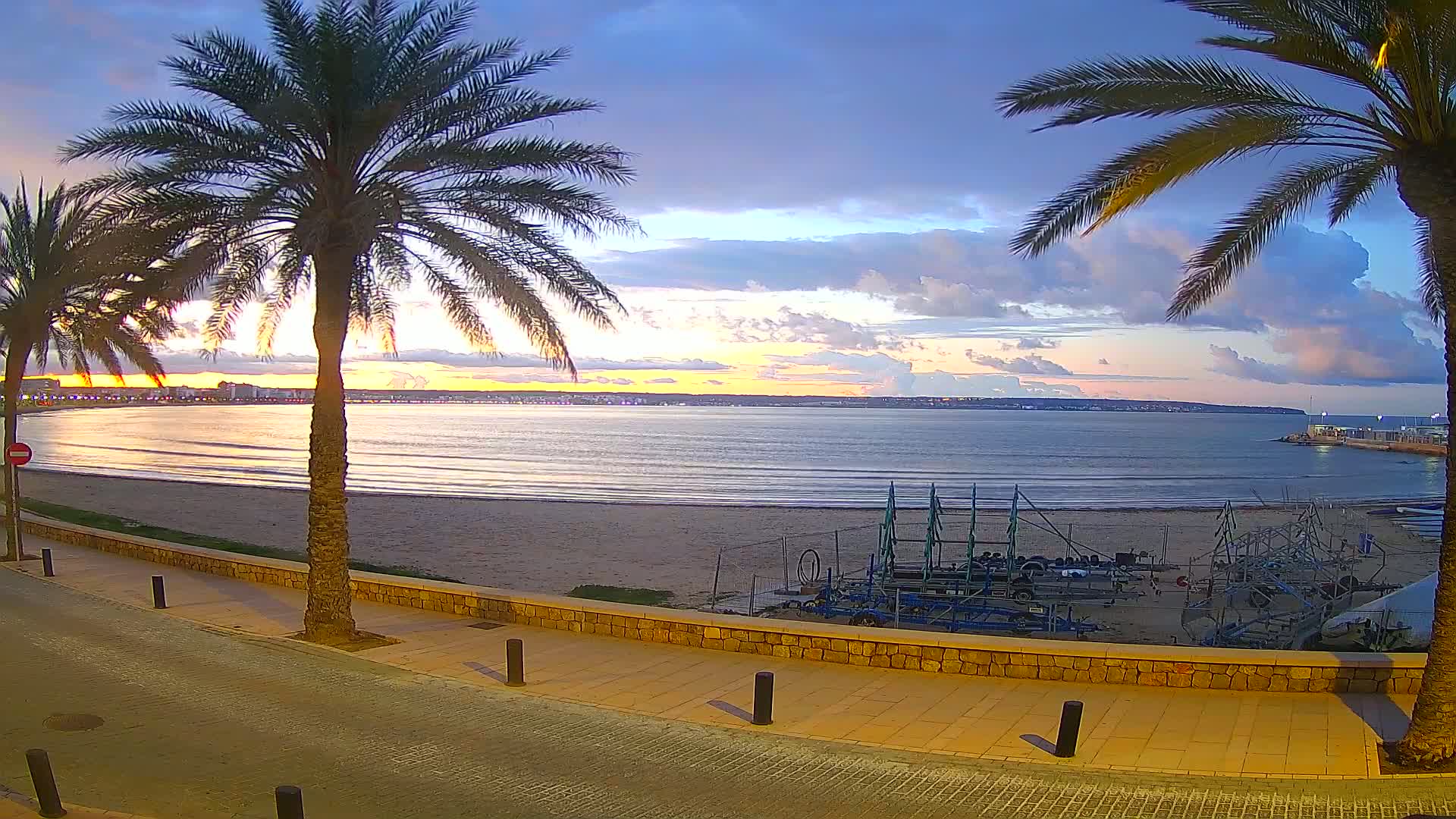 A sandy beach with calm waves, palm trees lining a paved walkway, and equipment stored on the sand, all under a partly cloudy sunset sky.
