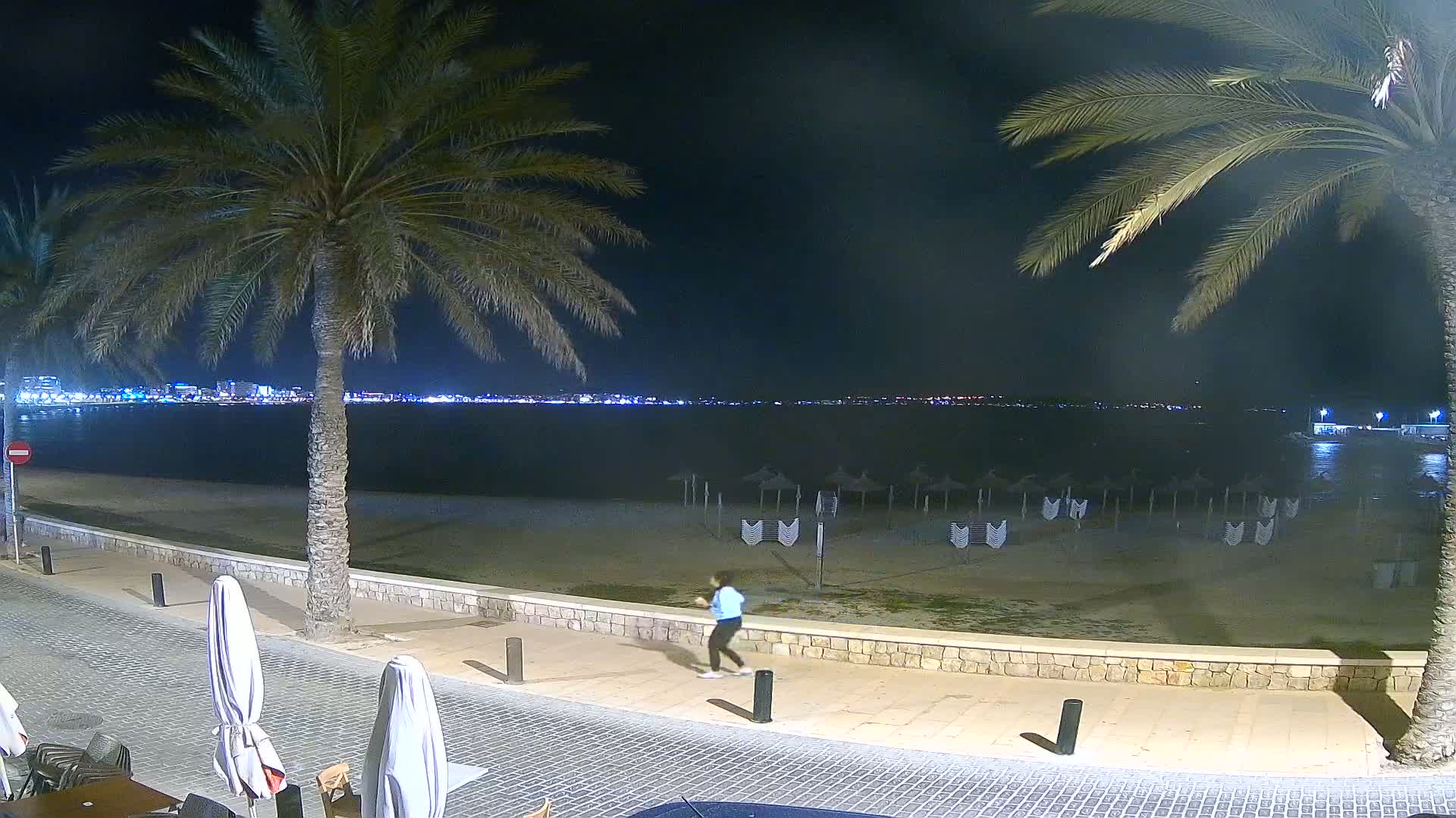 A person walks along a nighttime beachside promenade under a clear sky, with palm trees, a calm sea, and a distant, illuminated cityscape visible.