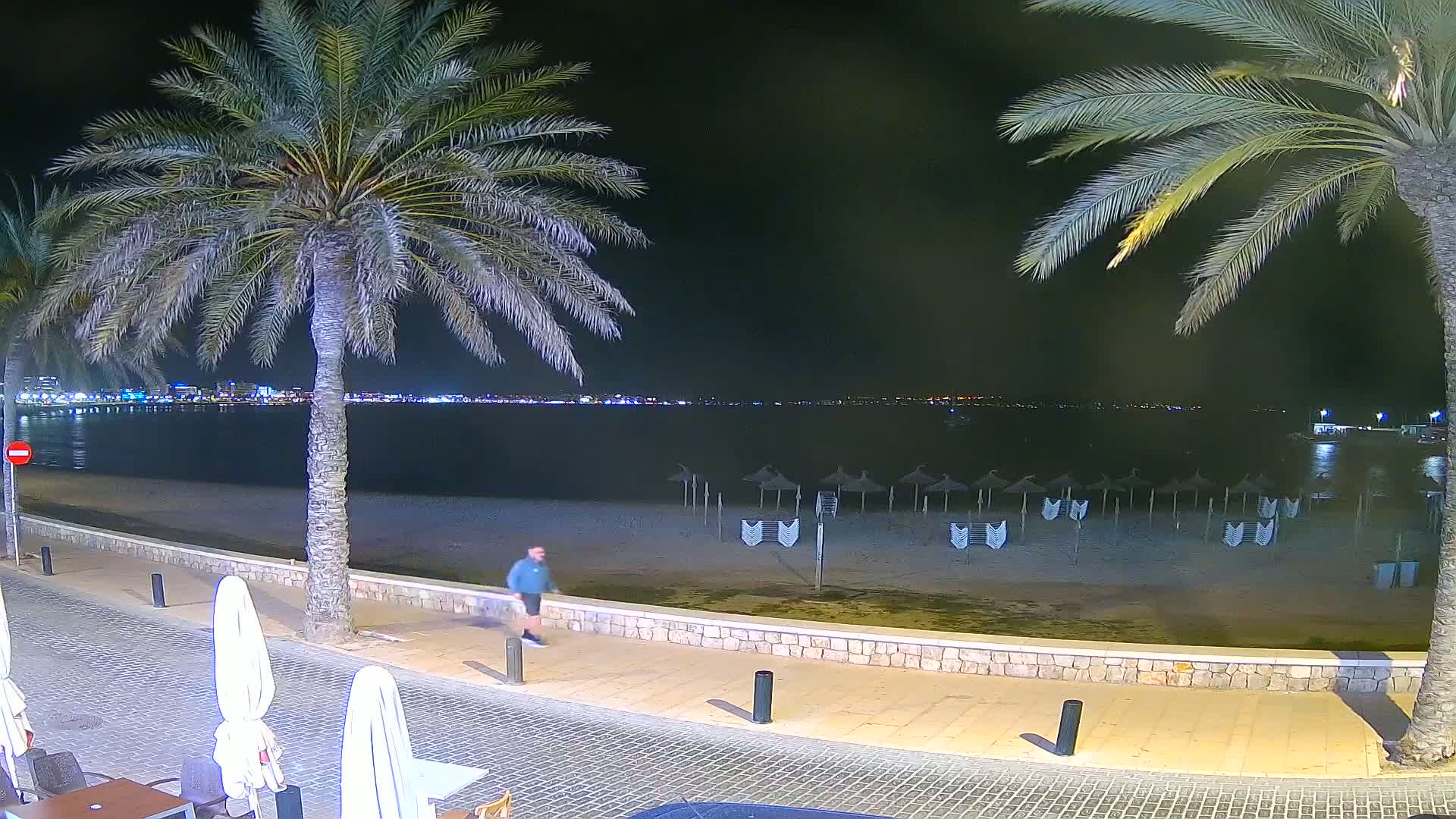 A man walks along a nighttime beach promenade, past closed beach umbrellas, toward a calm, city-lit ocean.