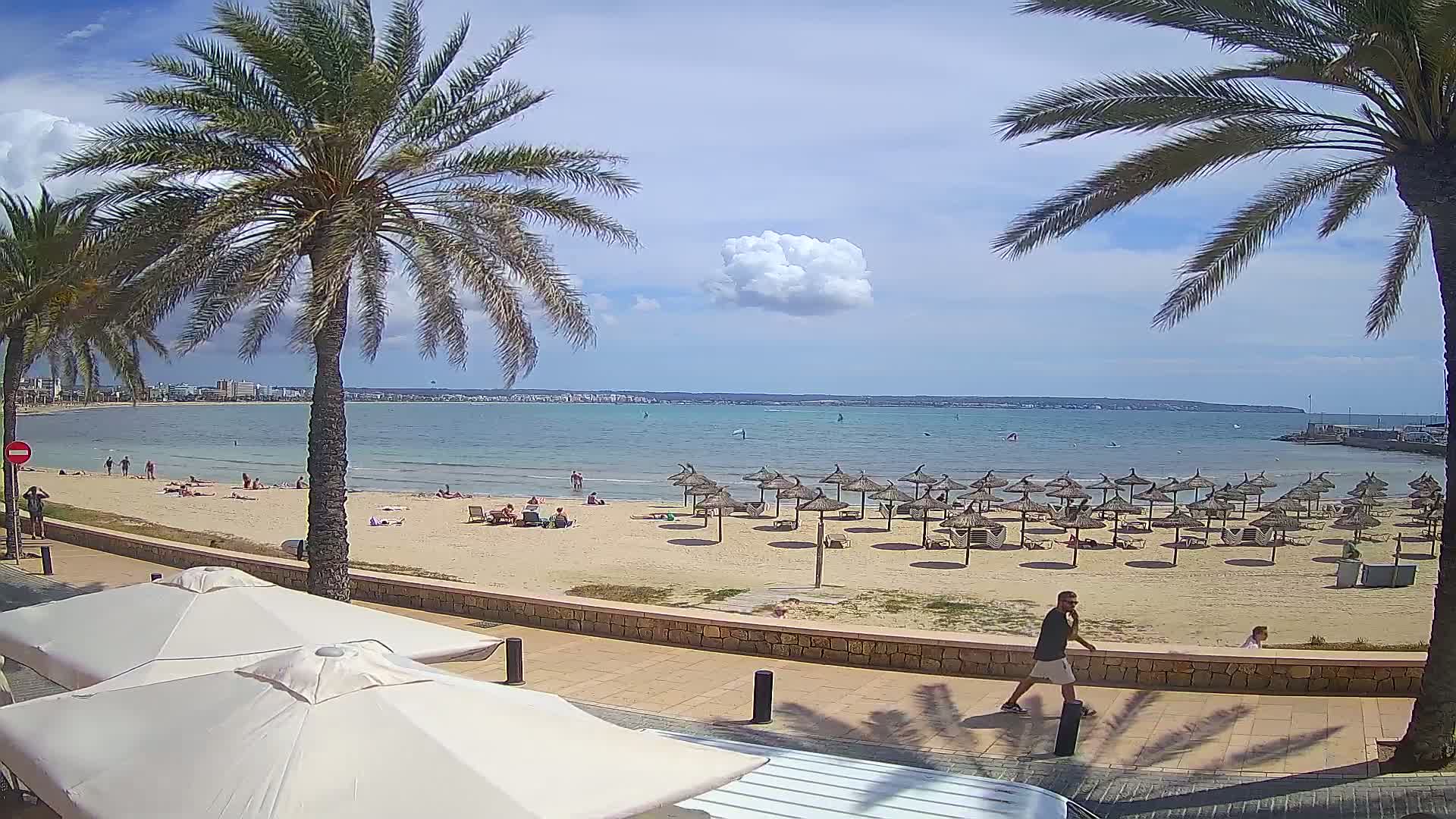 A sandy beach with numerous thatched umbrellas and scattered sunbathers is situated beside a calm, light-blue sea under a partly cloudy sky, palm trees framing the view.