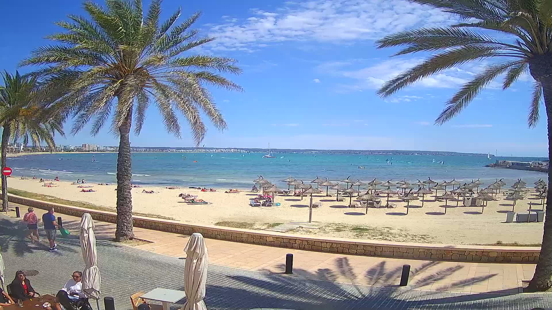 A sunny beach scene shows a sandy shore with numerous thatched umbrellas, people sunbathing and swimming in the calm, blue water, and palm trees framing the view.