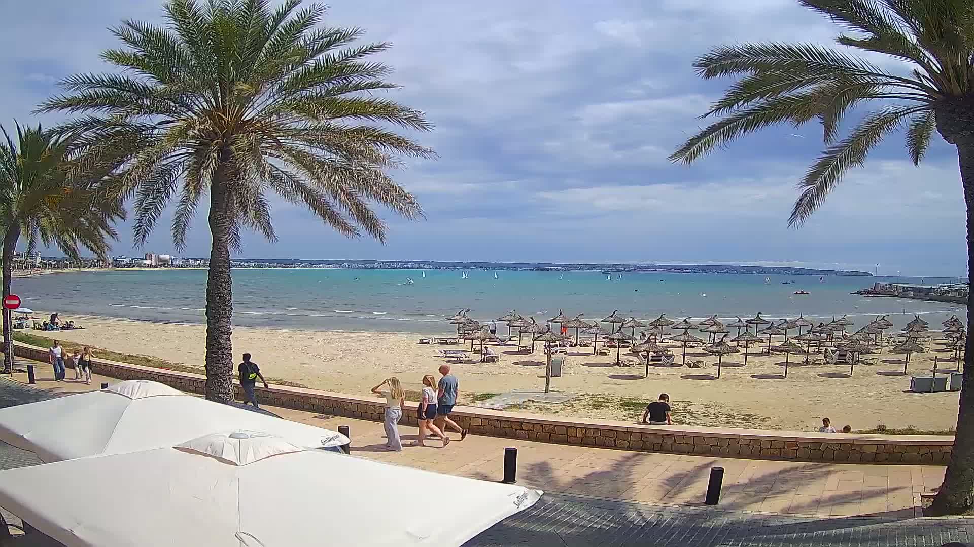 A sandy beach with numerous thatched umbrellas, palm trees, and people walking along a promenade overlooks a calm, light-blue bay dotted with sailboats under a partly cloudy sky.
