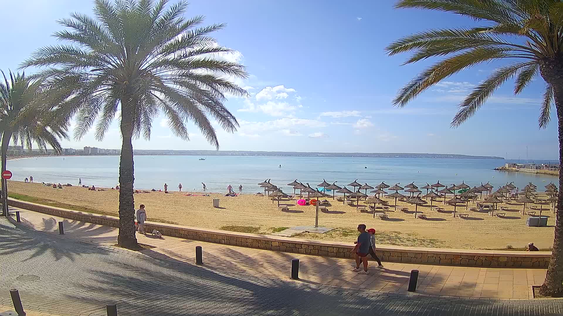 A sandy beach with calm, clear water, numerous straw umbrellas, and scattered beachgoers is viewed from a paved walkway lined with palm trees under a mostly sunny sky.