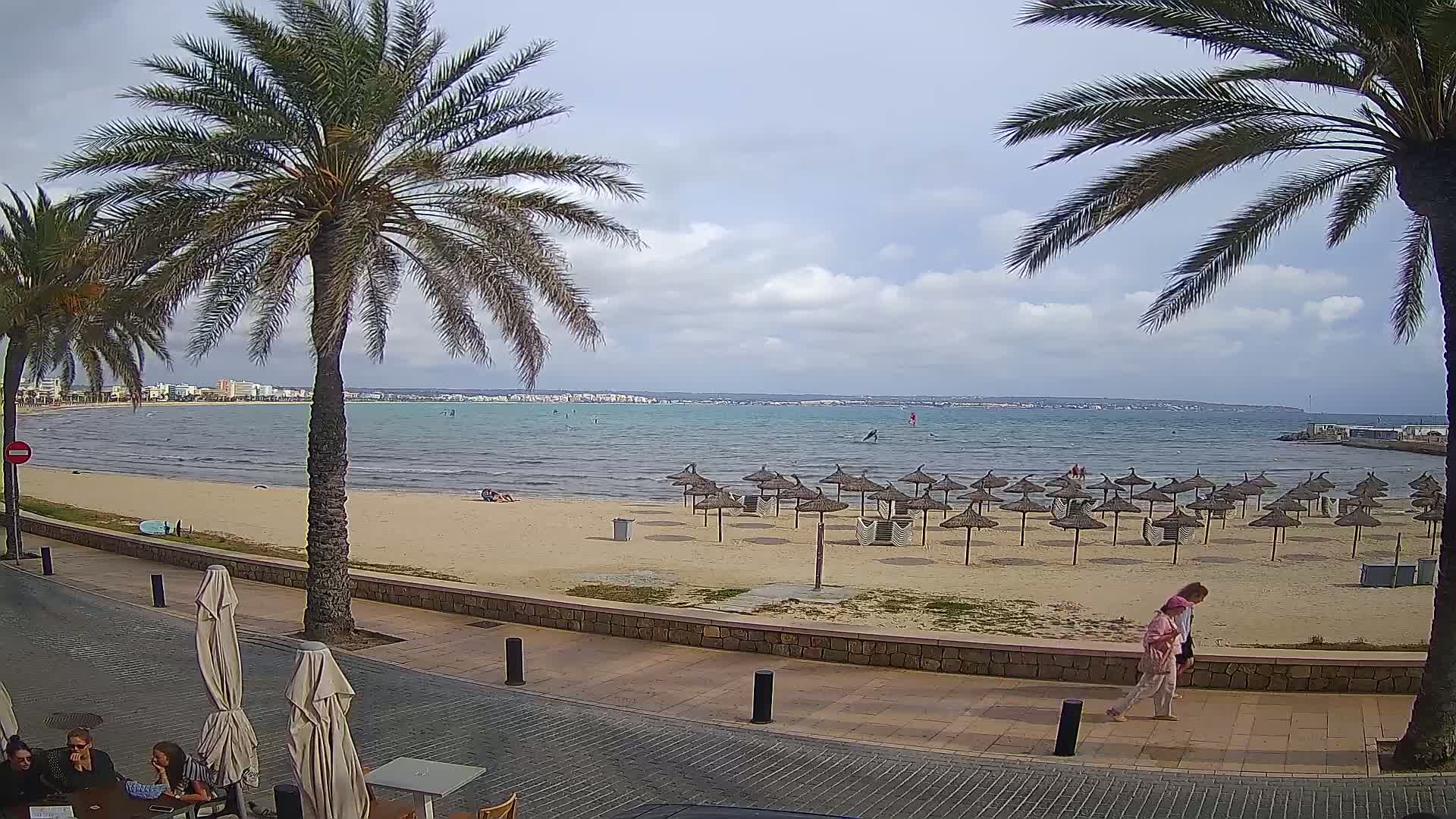 A sandy beach with numerous straw umbrellas stretches along a calm, blue-green sea under a partly cloudy sky, with palm trees lining a paved walkway in the foreground.
