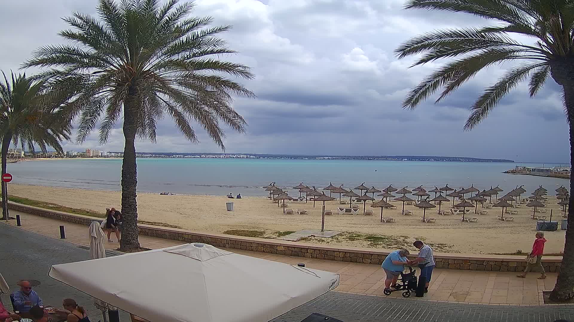 A sandy beach with numerous straw umbrellas under a cloudy sky, palm trees, and people walking along a promenade next to a calm, turquoise body of water.