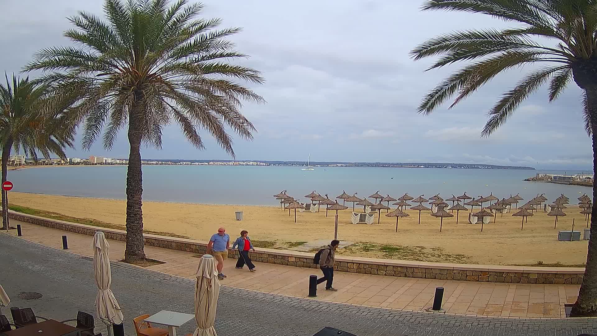 A sandy beach with numerous straw umbrellas, palm trees, and several people walking along a paved promenade overlooking a calm bay under an overcast sky.
