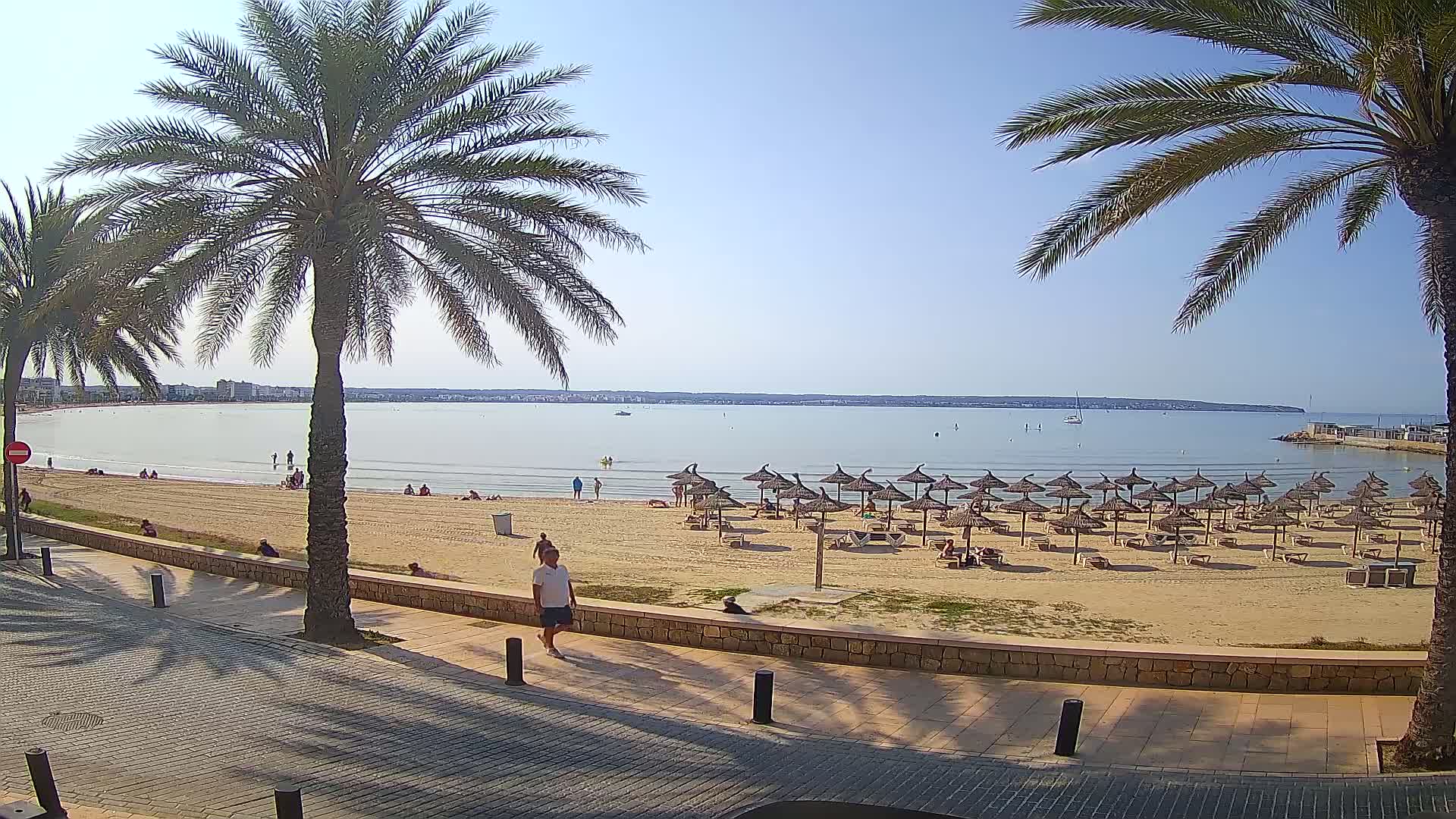 A sandy beach with many straw umbrellas and people is visible across a calm bay on a sunny day, viewed from a paved walkway lined with palm trees.