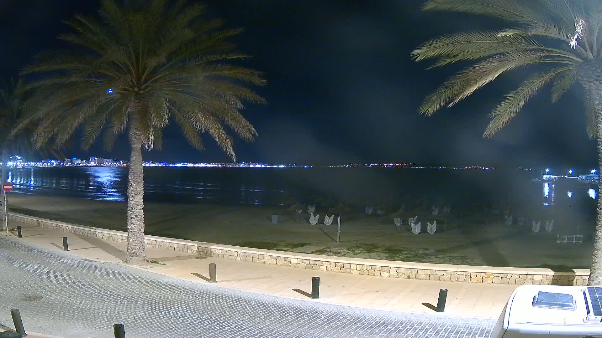 At night, a sandy beach with folded lounge chairs is visible, bordered by a paved walkway and palm trees, overlooking a calm body of water reflecting city lights in the distance.